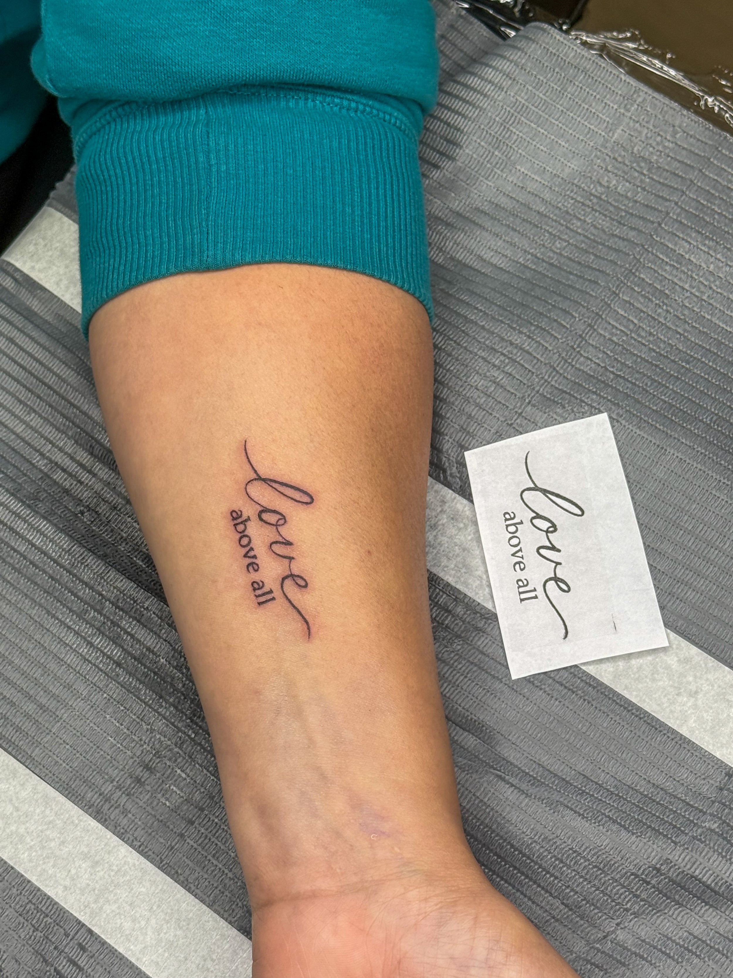 A tattoo on a person's forearm that reads, 'Love above all' in cursive script.
