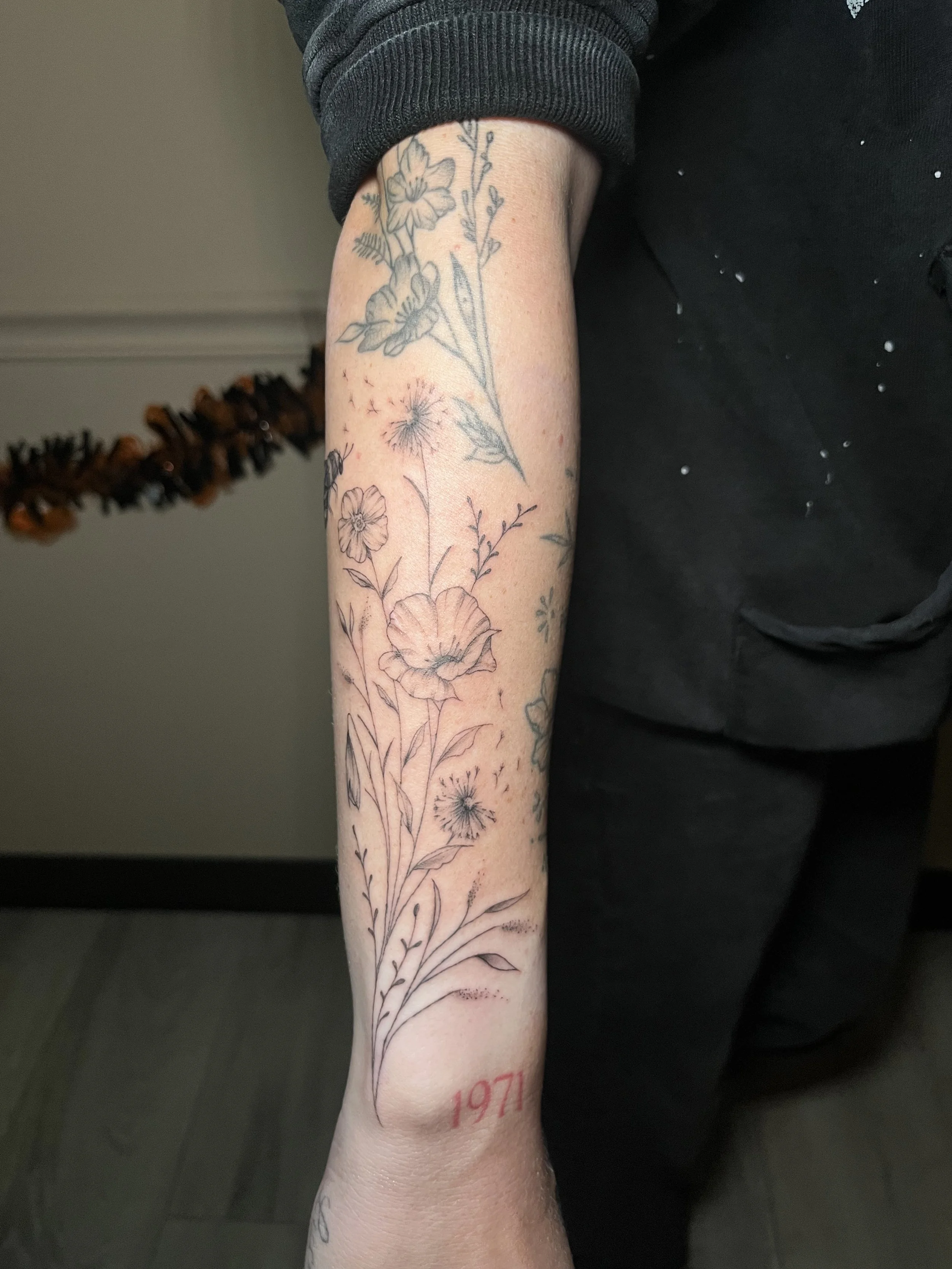A person's forearm with a floral tattoo design and the year "1971" tattooed in red near the wrist. The person is wearing a black shirt with splatters and a black jacket sleeve rolled up.