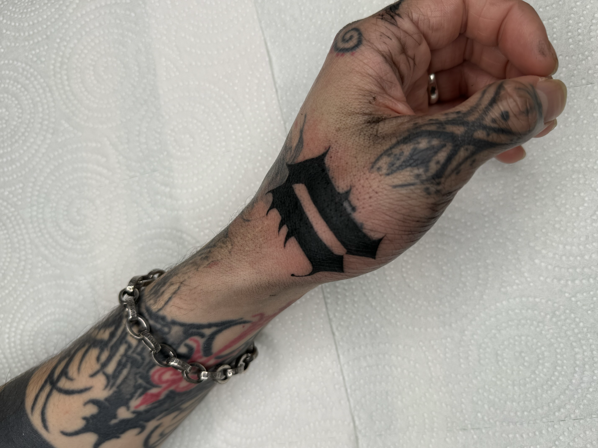 Tattooed hand with a chain bracelet resting on a white textured surface.