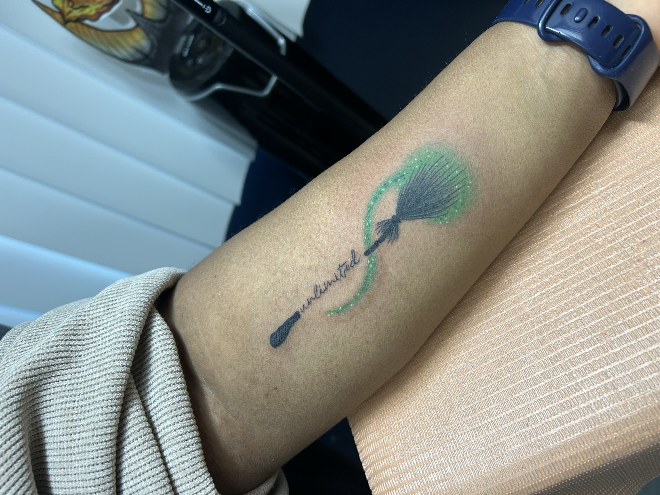 Tattoo of a makeup brush with the word 'unlimited' written below it, on a person's forearm.