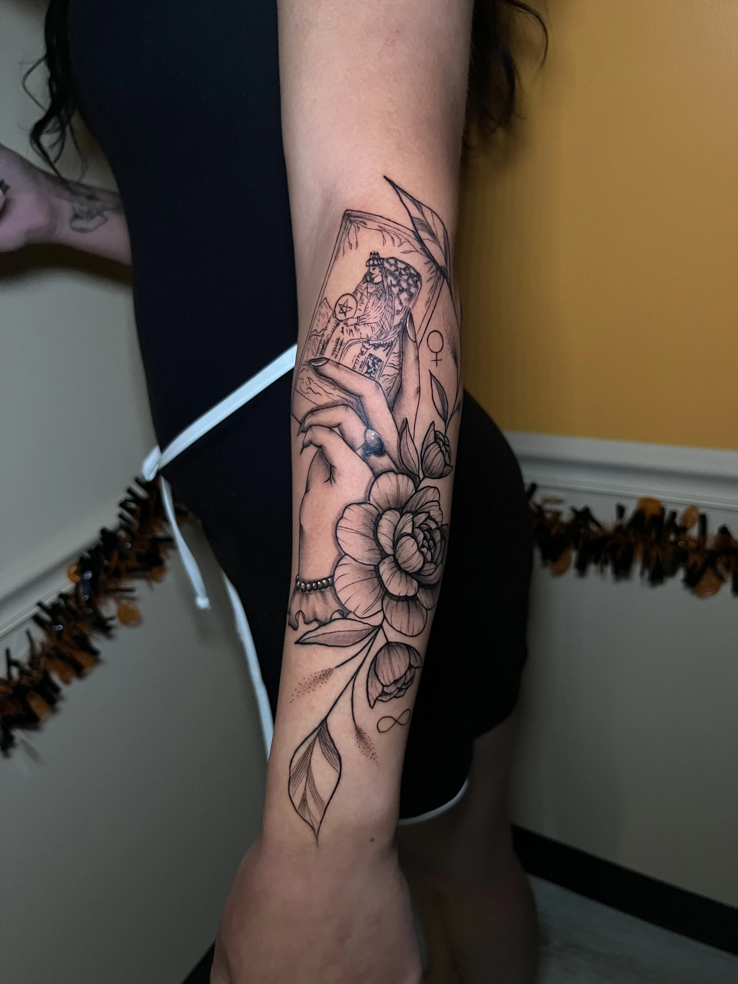 A person's forearm with a tattoo of a tarot card, a large flower, and a hand with a gemstone ring.