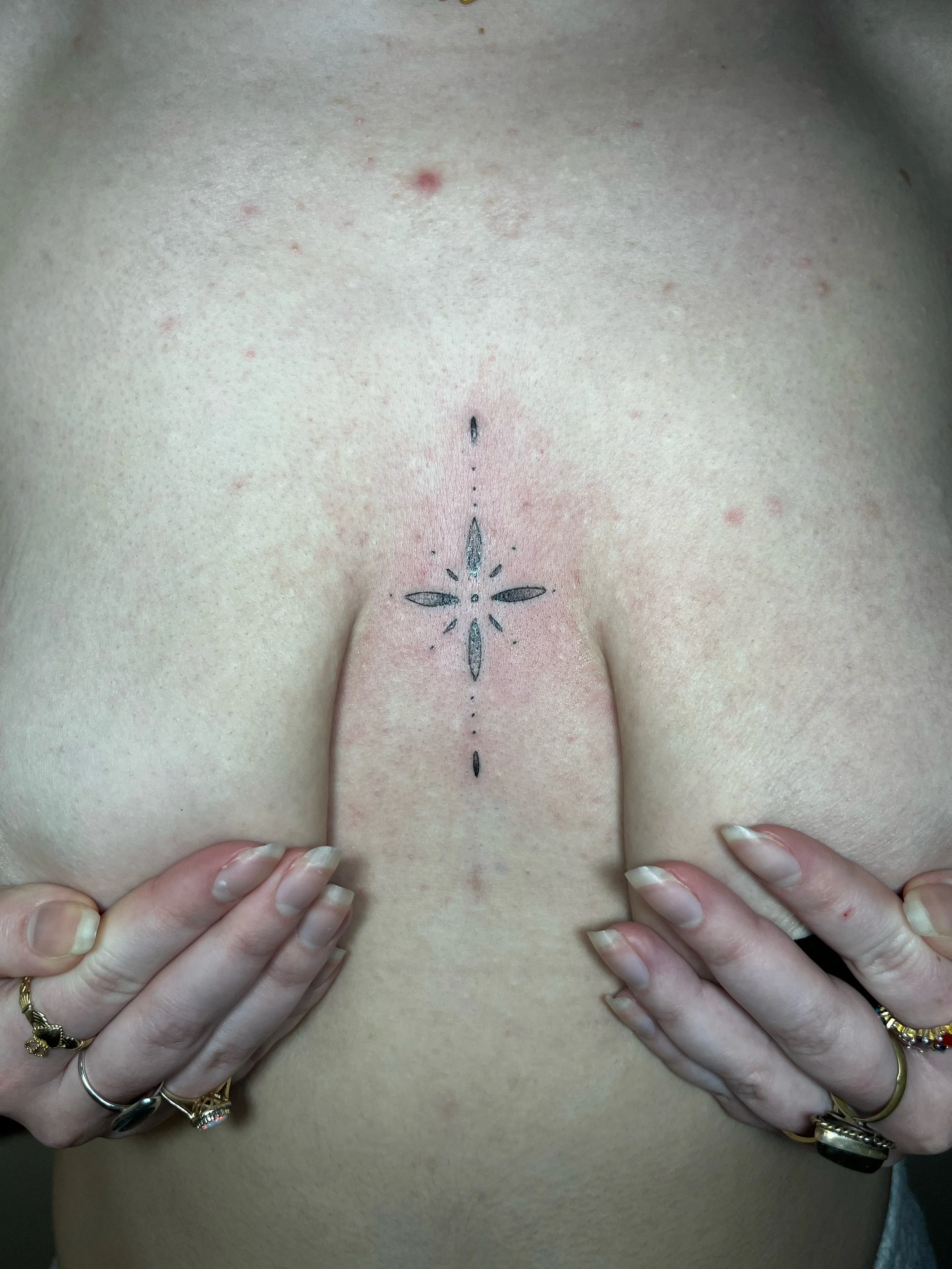 A close-up of a person's chest with a decorative black tattoo in the center, surrounded by multiple rings on their fingers, with hands holding and slightly pulling their breasts apart.