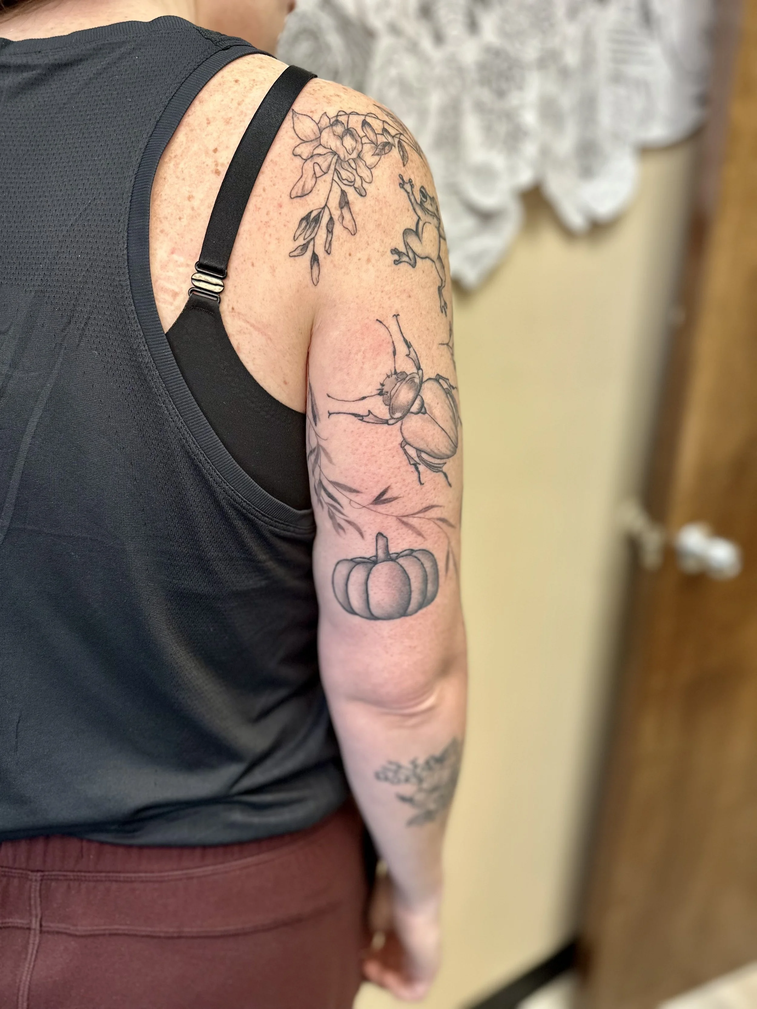 Close-up of a person's upper arm and shoulder with tattoos of a flower, a frog, a pumpkin, and a bee, wearing a black sleeveless top and maroon pants.