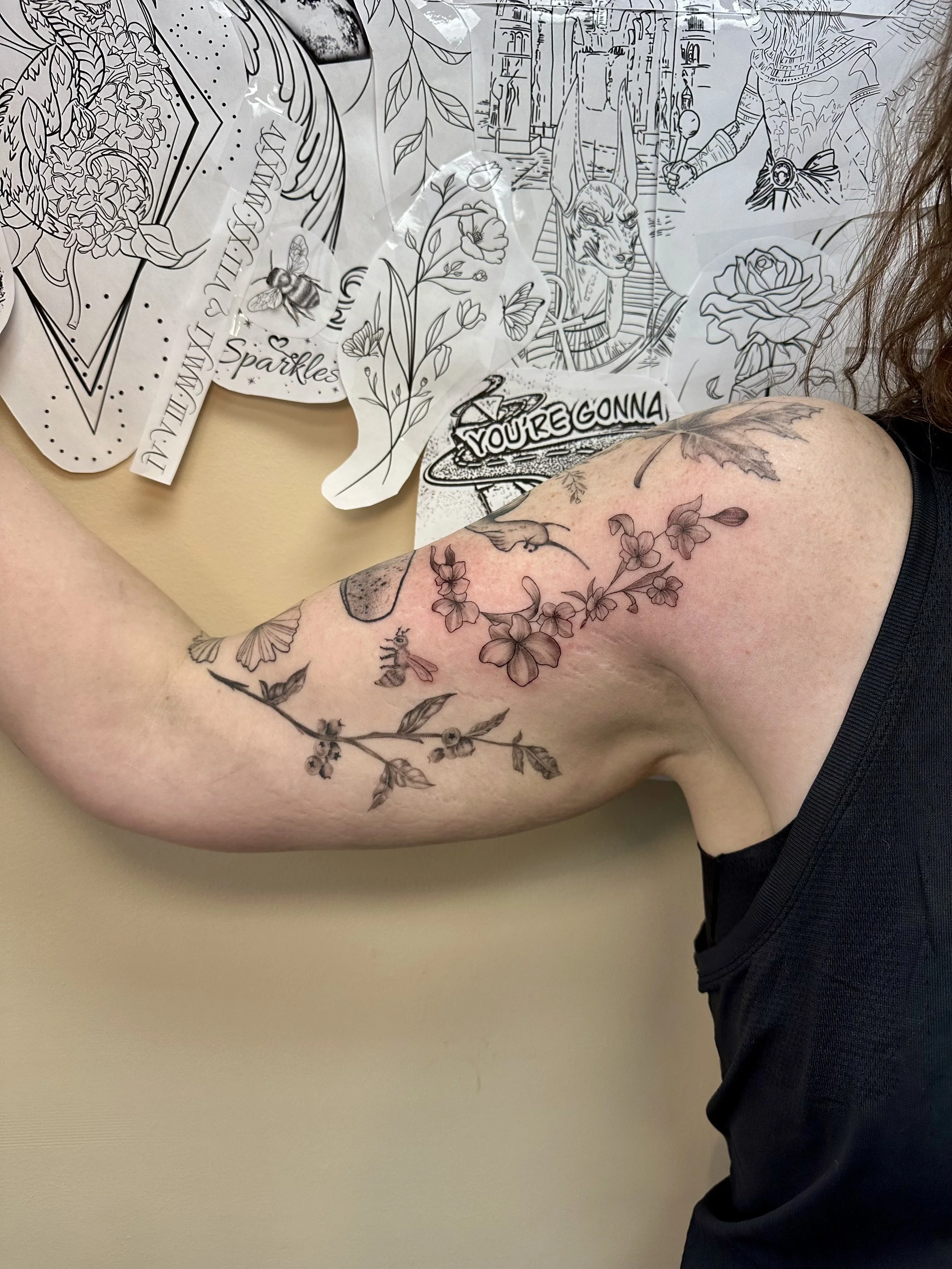 Close-up of a person's upper arm with a floral tattoo featuring cherry blossoms and leaves. The person is wearing a black sleeveless shirt, and the background includes wall art, stickers, and drawings.
