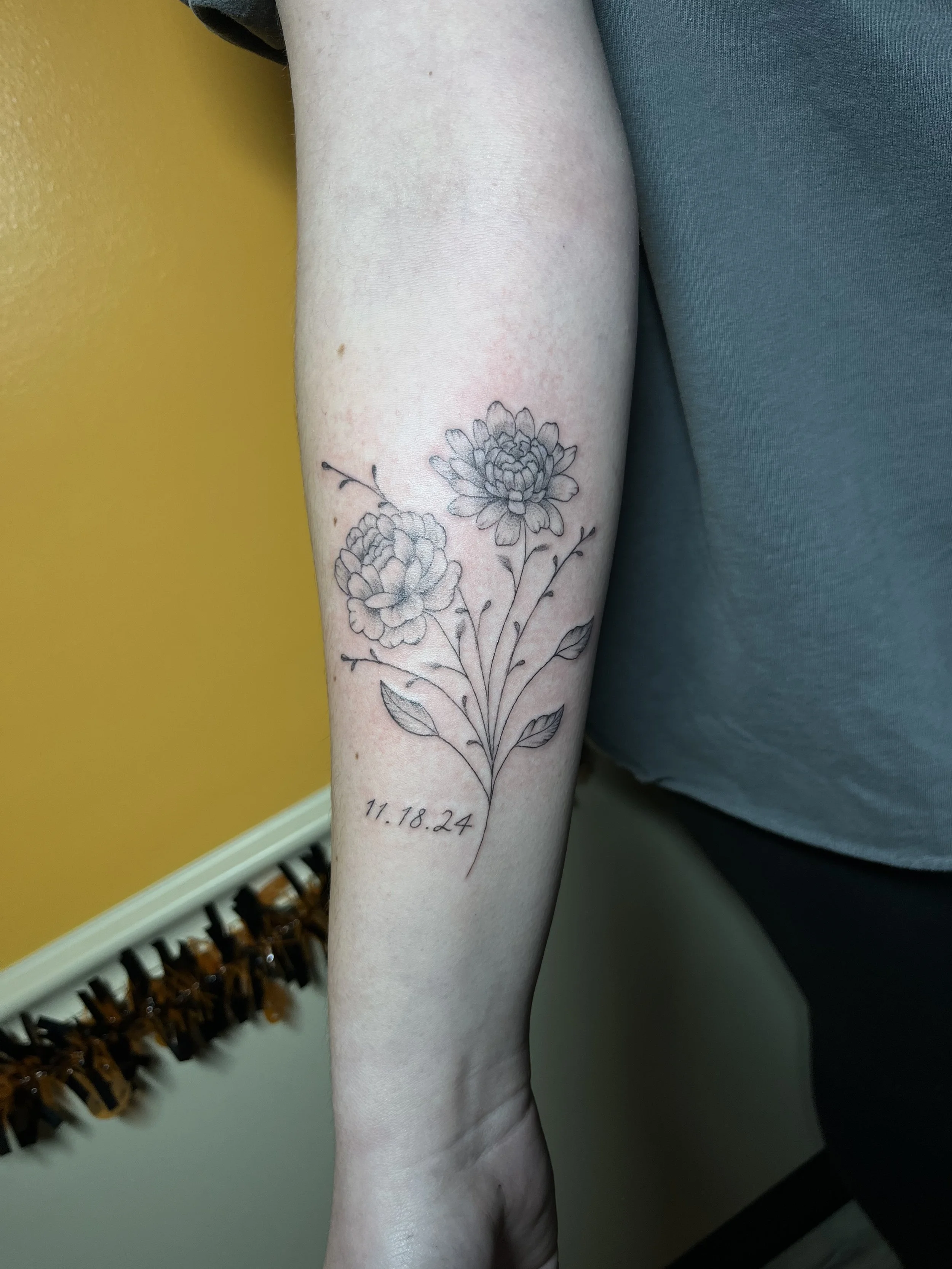 Black and gray floral tattoo on inner forearm with a date '11.18.24' below the flowers.