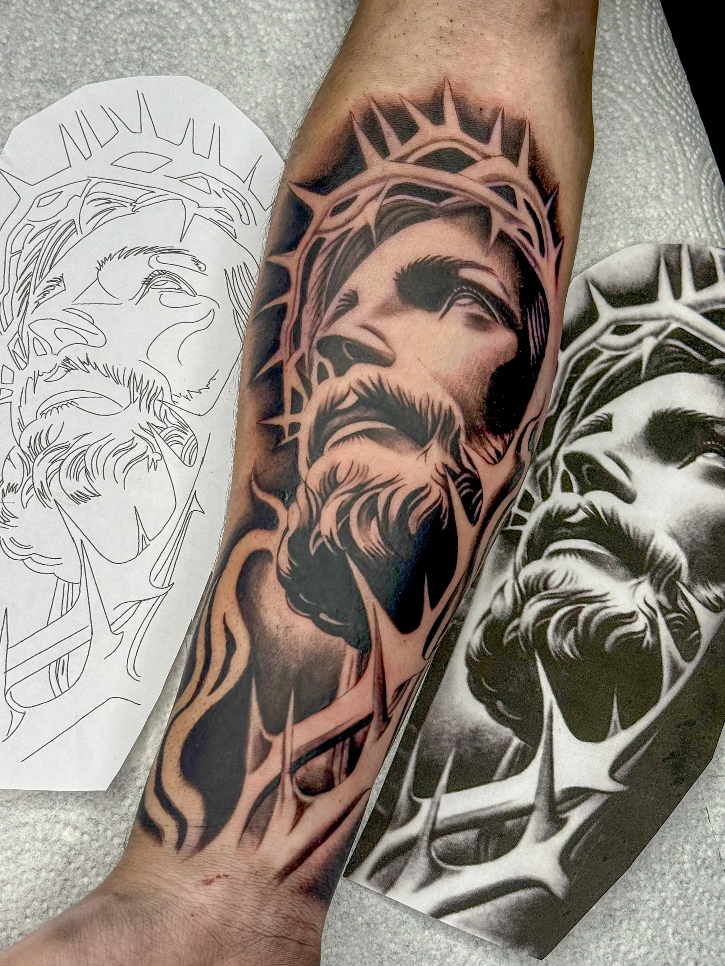 A tattoo of a bearded man's face surrounded by a halo, created with black ink and shades of pink, on a person's forearm. The tattoo is inspired by religious iconography, with detailed shading and bold lines.