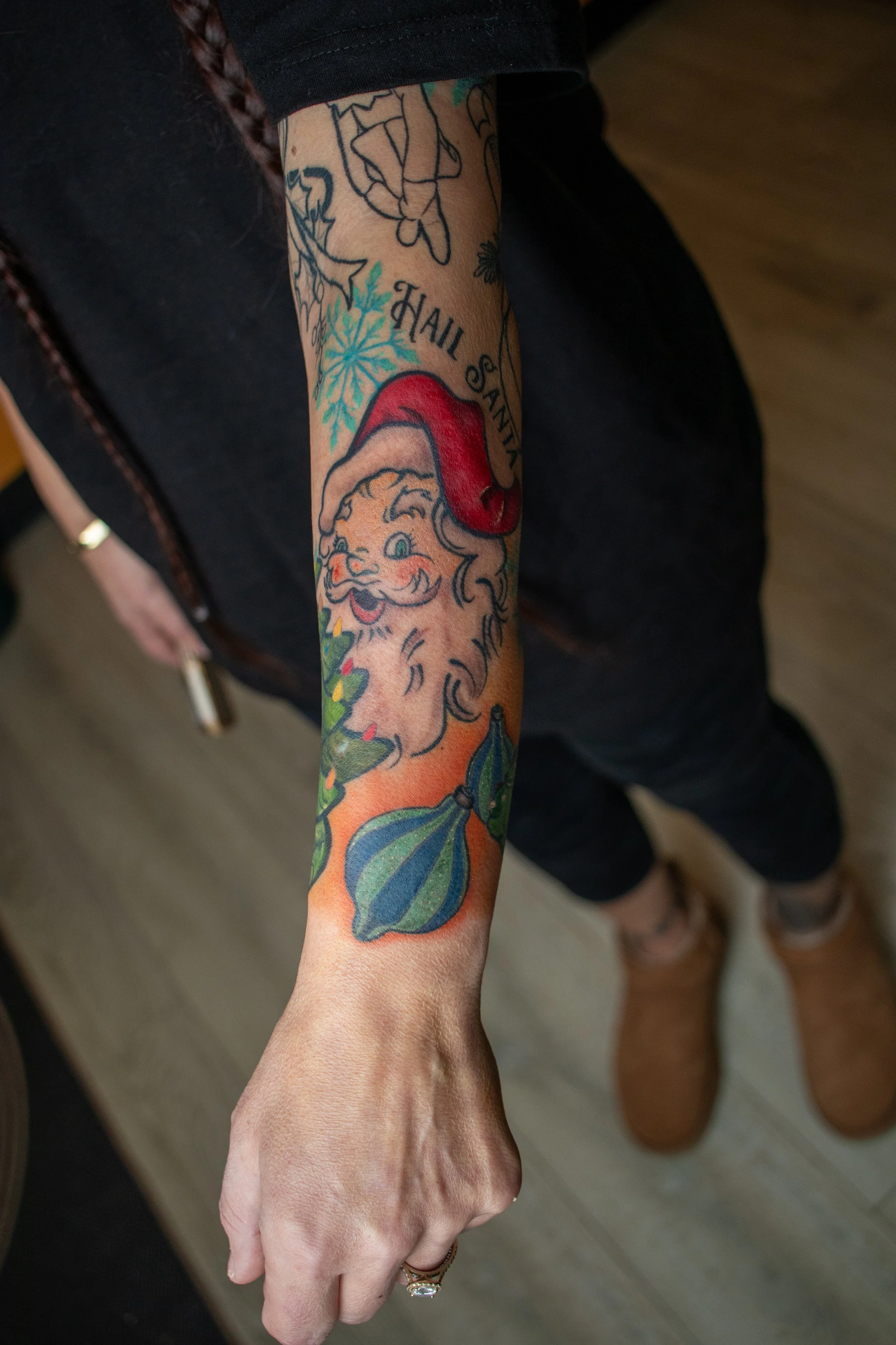 Colorful tattoo of Santa Claus's face with a red hat, green and purple striped Christmas ornaments, holly, and a snowflake, with the text 'Hail Santa' on the arm.