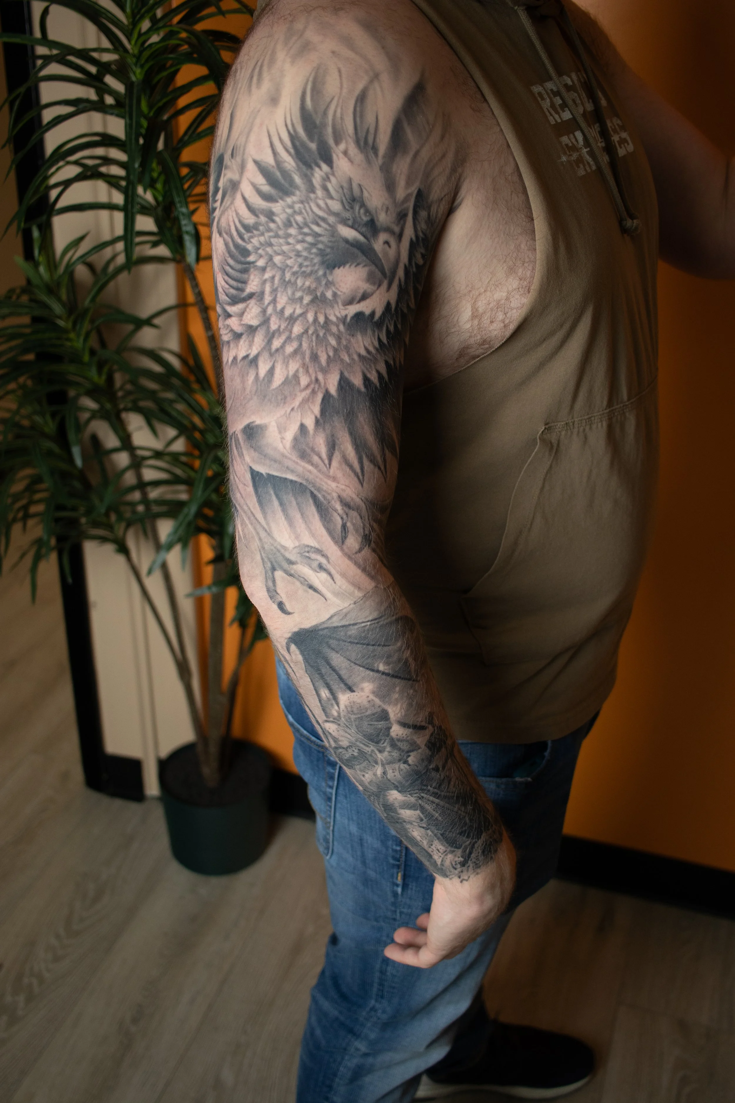 A person with a detailed black and gray tattoo sleeve of an eagle and a dragon on their arm, wearing a sleeveless shirt and jeans, standing indoors near a green potted plant.