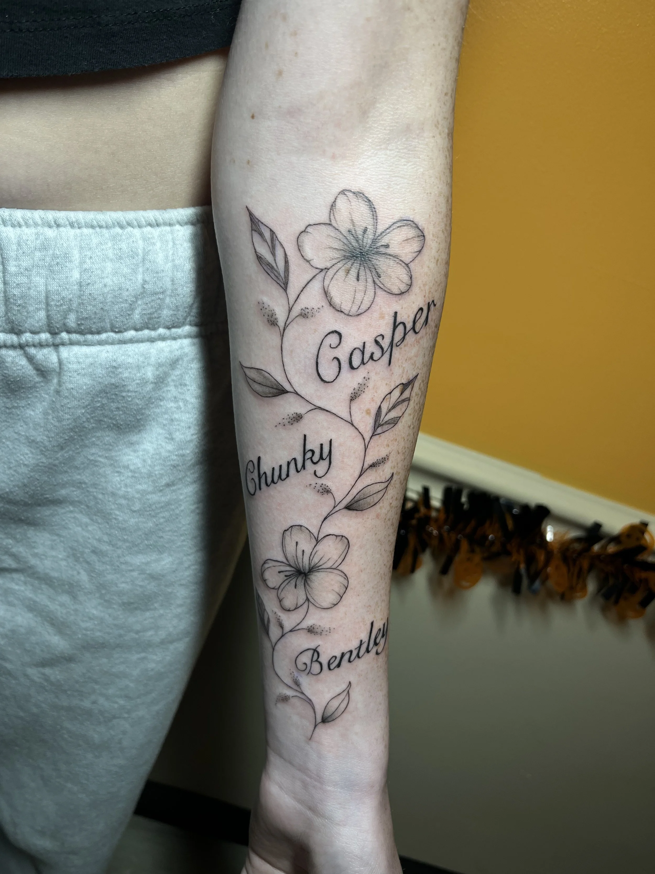 A forearm tattoo of a flower with detailed petals and leaves, with the names 'Gasper,' 'Chunky,' and 'Bentley' written in cursive script.