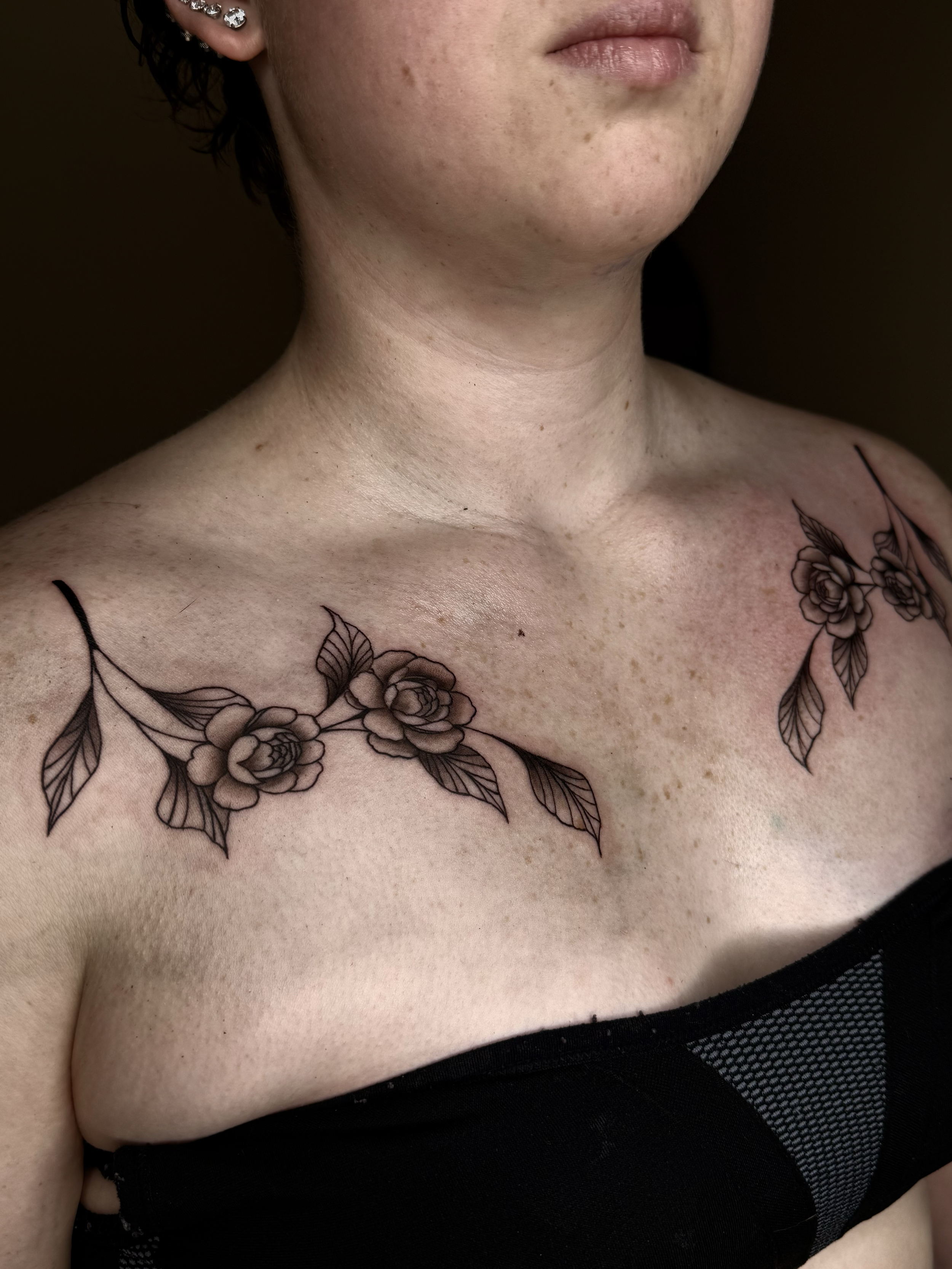 Close-up of a woman's shoulder and upper chest displaying a floral tattoo with roses and leaves on both sides of her collarbone.
