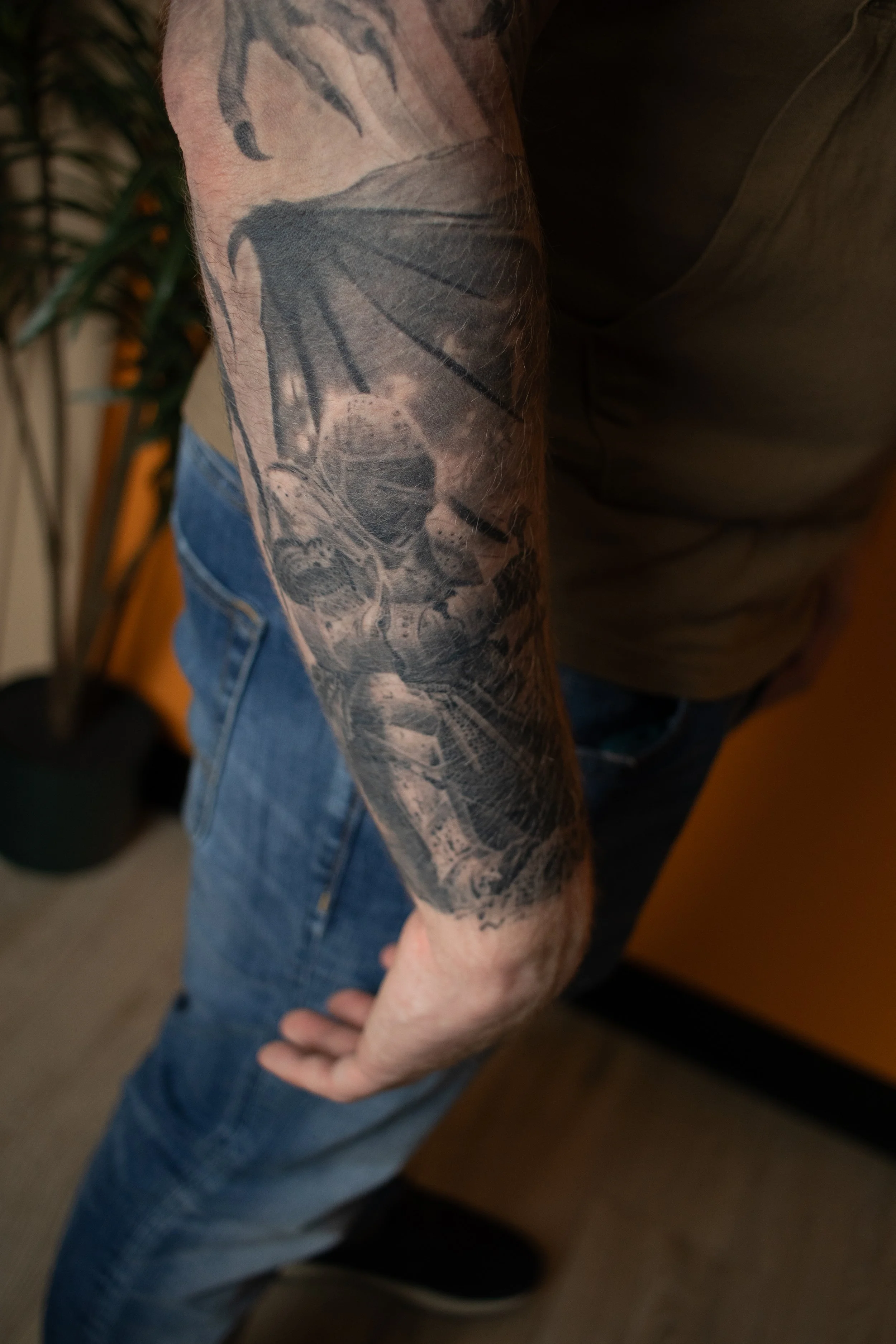 A person with a tattooed arm wearing a dark T-shirt and blue jeans, standing indoors near a plant and a yellow object.
