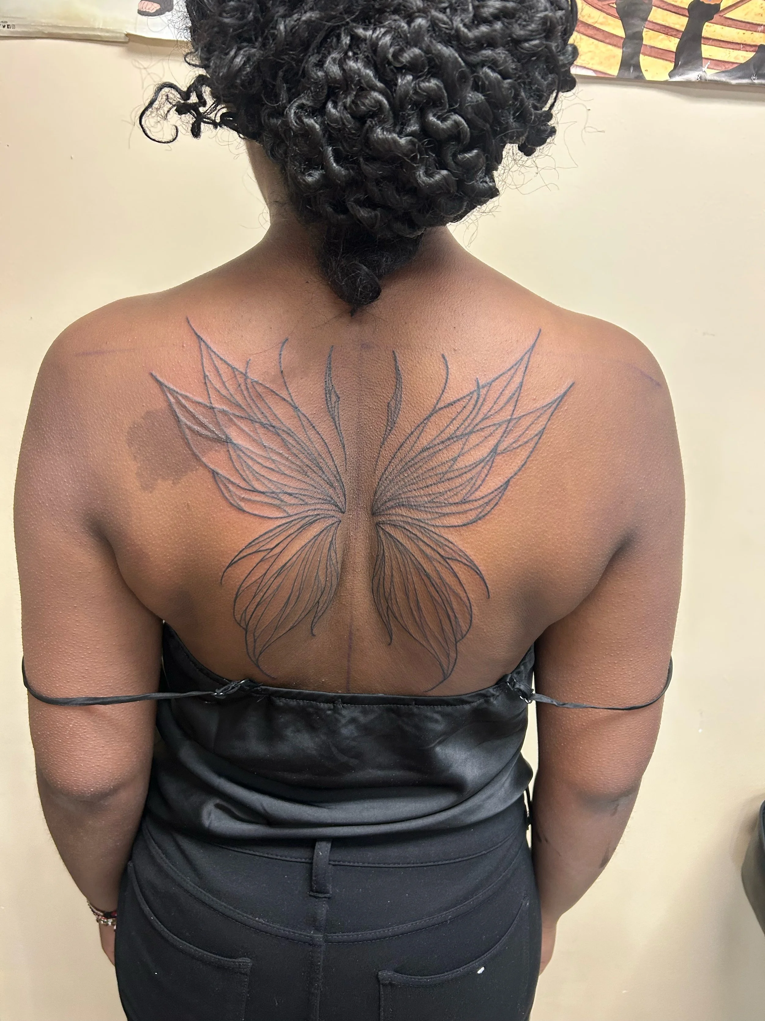 Back of a woman with curly hair and a butterfly tattoo on her upper back, wearing a black top and black pants.