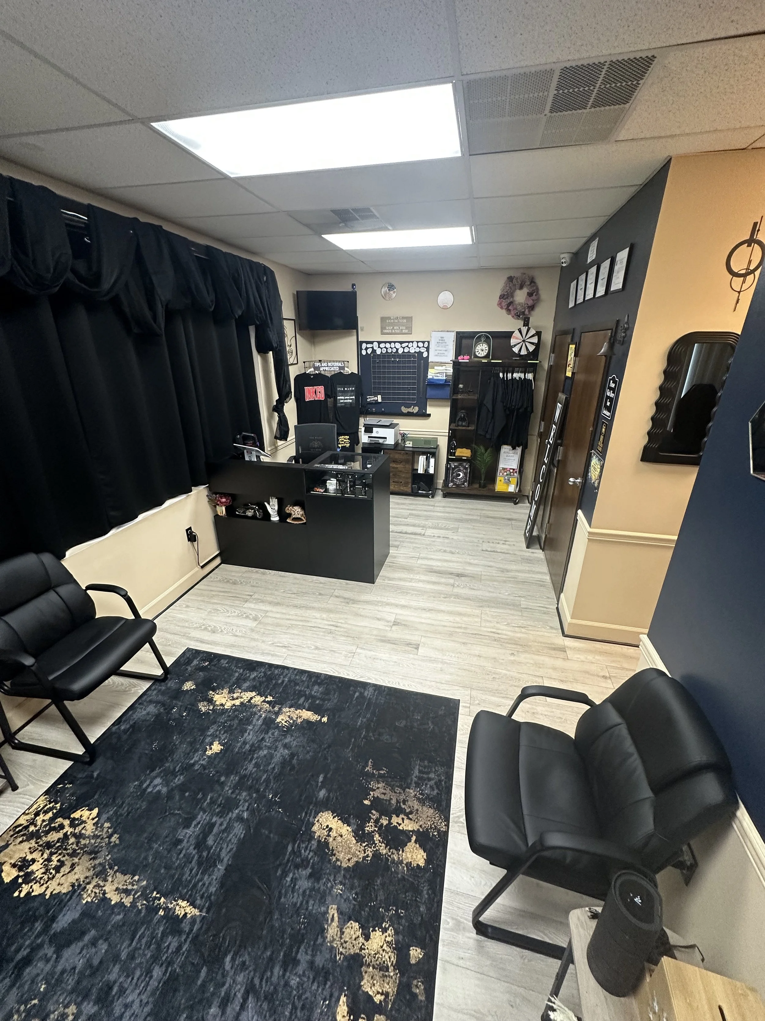 A room with black curtains, seating chairs, a black rug, and wall decorations including a dartboard, framed pictures, and a wreath. There are office or retail items on desks and shelves.