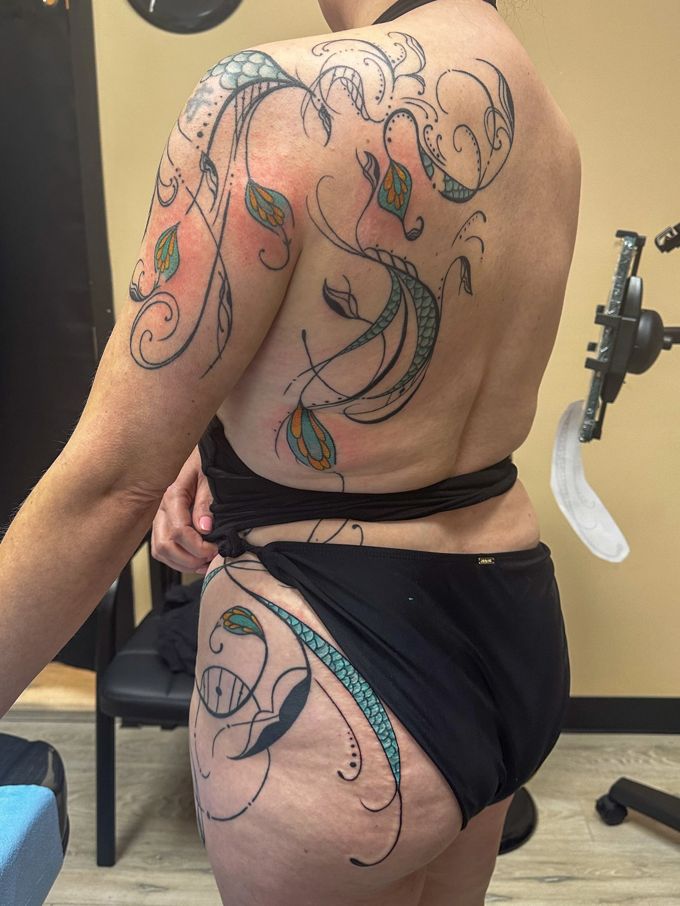 A person with a large, colorful tattoo of a dragon on their back and thigh, sitting in a gym setting.