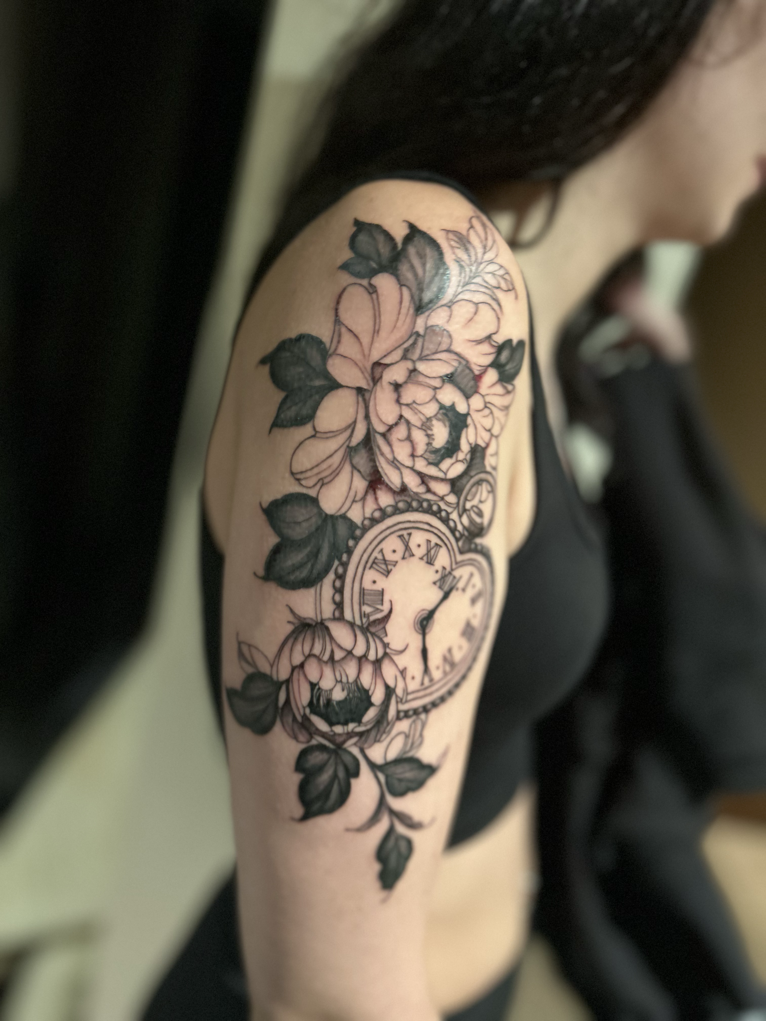 Close-up of a person's shoulder and upper arm with a black and gray floral tattoo featuring large flowers and leaves surrounding a clock face with Roman numerals.