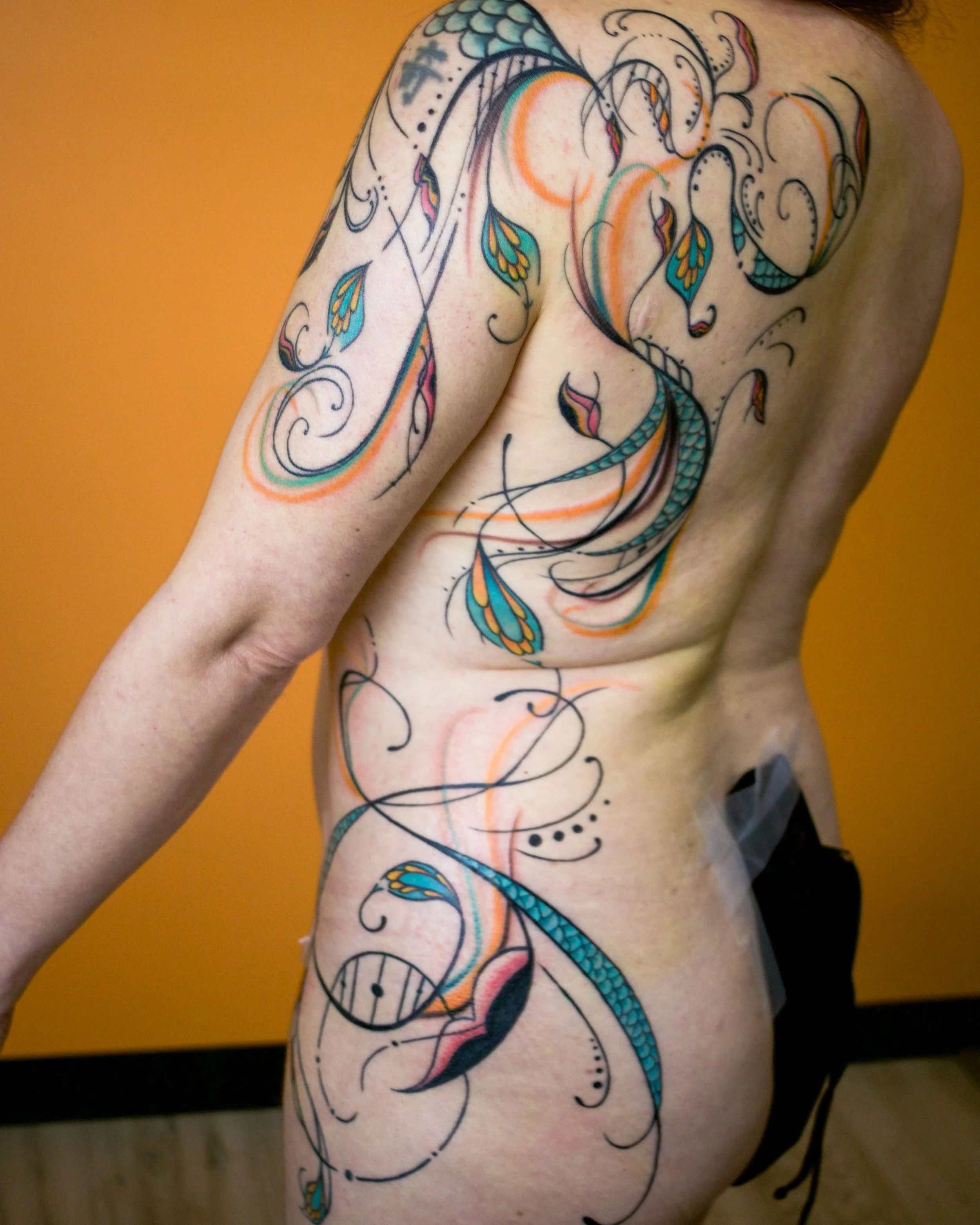 Person with colorful tattoo artwork on back and arm, featuring a snake, leaves, and abstract swirls in black, orange, teal, and pink ink.