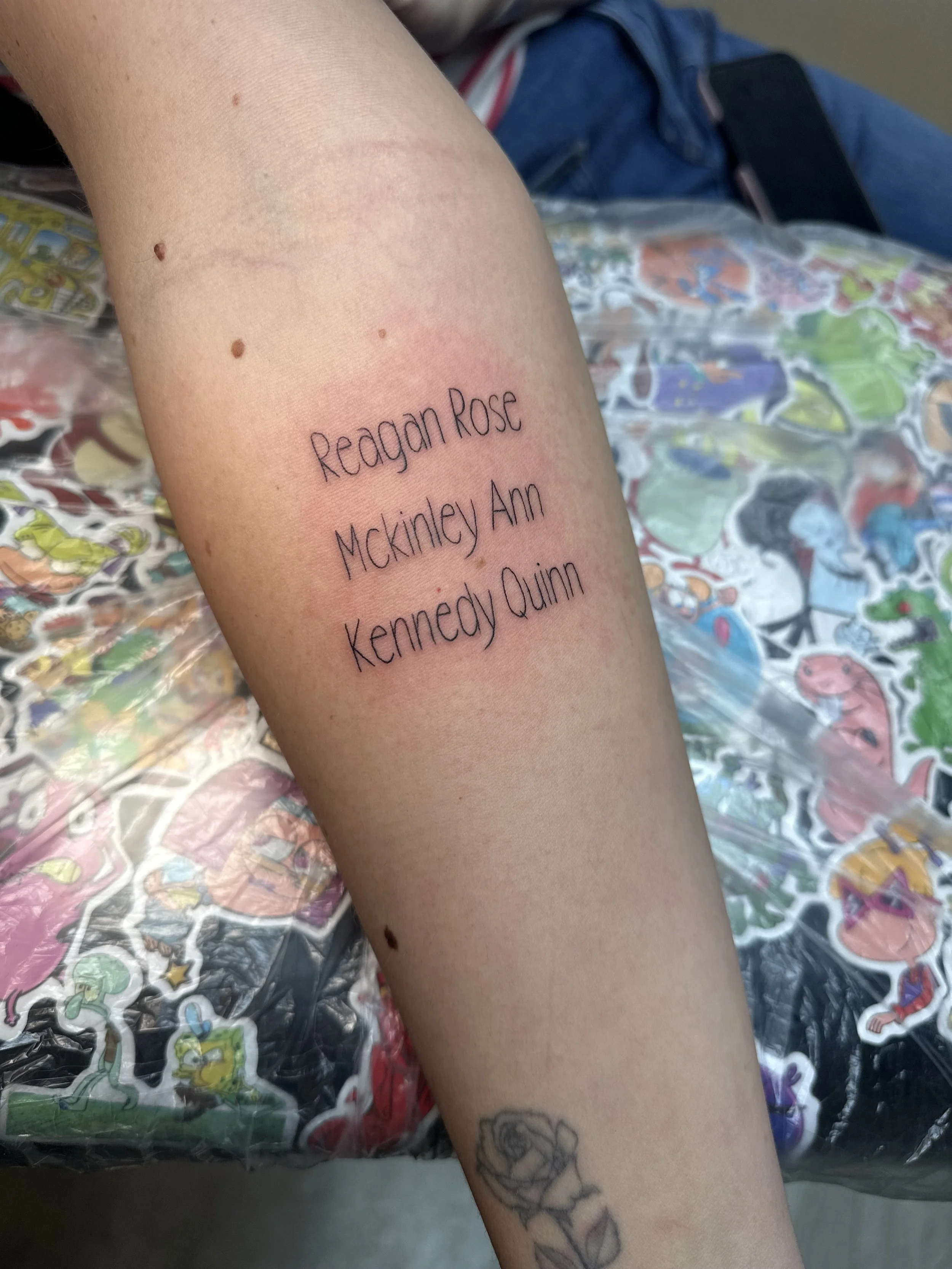 Tattoo on someone's forearm with names 'Reagan Rose', 'Mckinley Ann', and 'Kenneedy Quinn' in black ink, above a line drawing of a rose. The arm is resting on a surface covered with stickers.