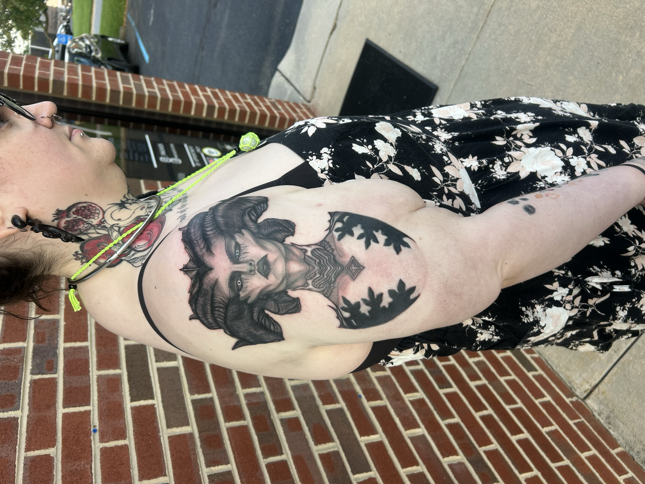 A woman with tattoos on her shoulder and neck, wearing black and white floral clothing, standing outside next to a brick wall and a parking lot.