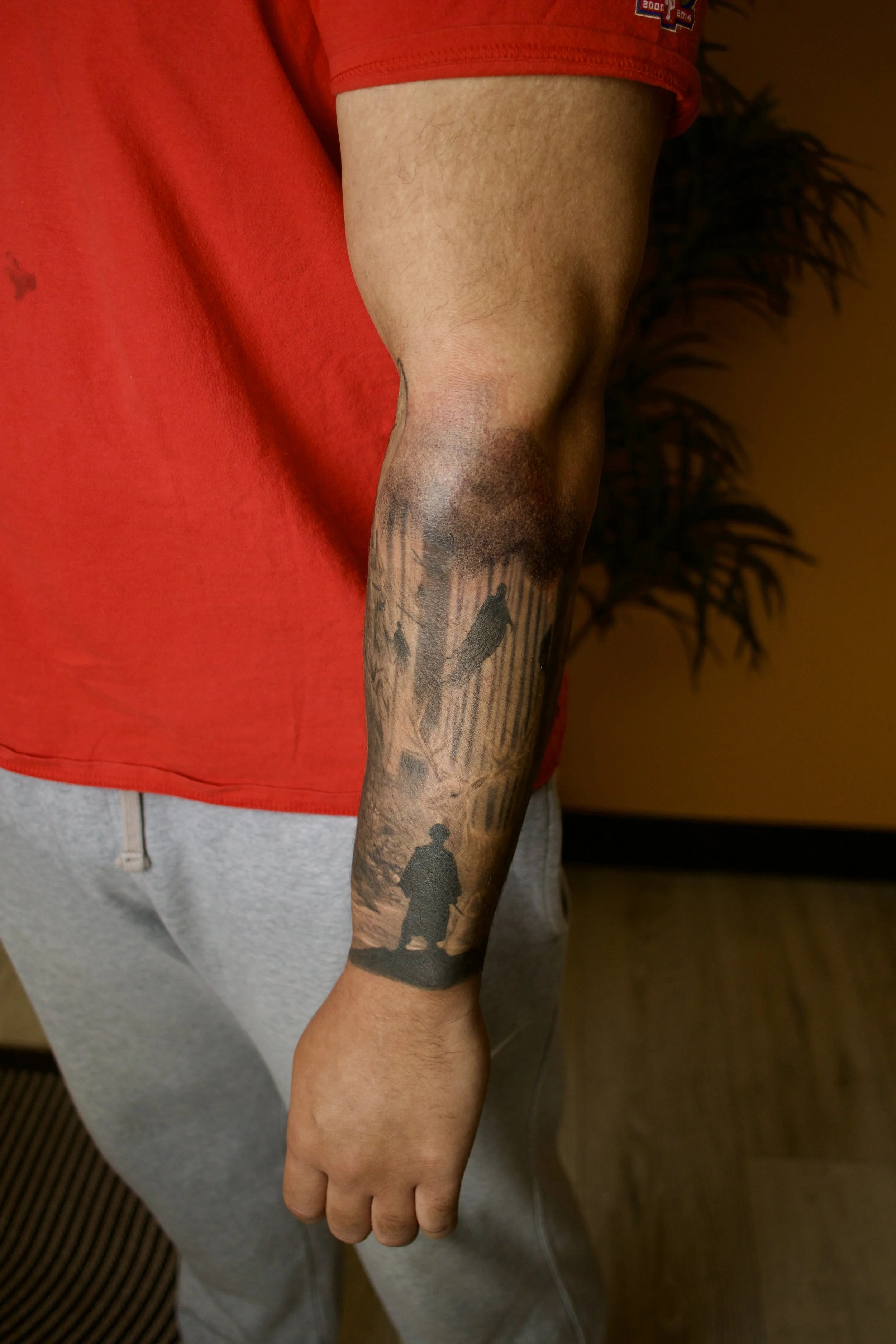 Close-up of a man's right arm with a tattoo of two silhouettes, one standing and one perched on a branch, set against a background of trees and a pathway in a forest.