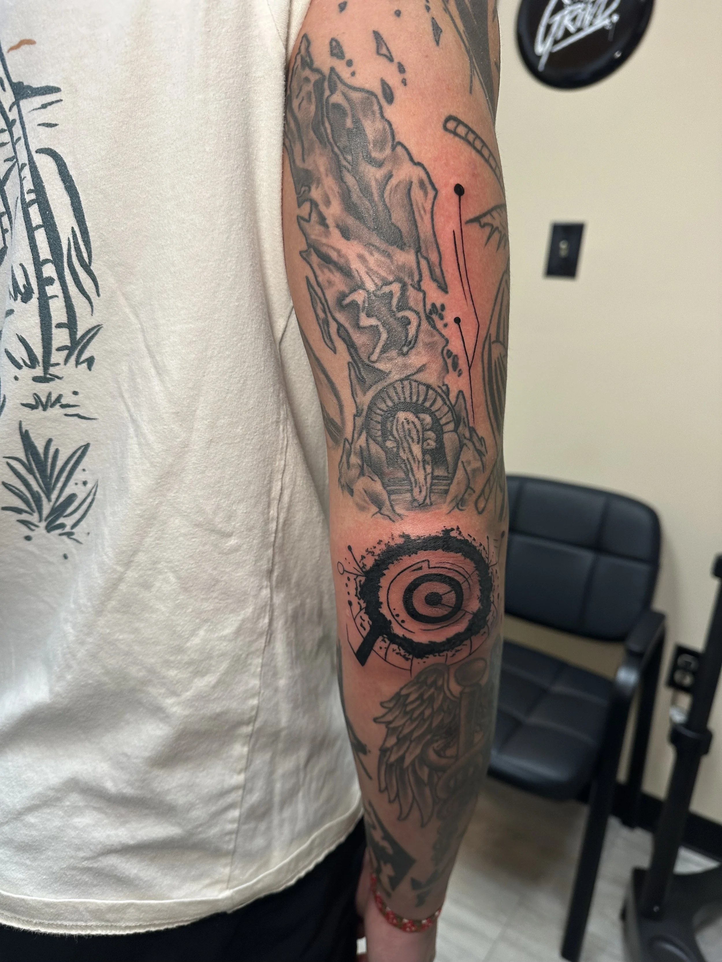 Tattoo sleeve on person's arm featuring mountainous landscape, a waterfall, an archway, a target symbol with a splatter effect, and various abstract designs