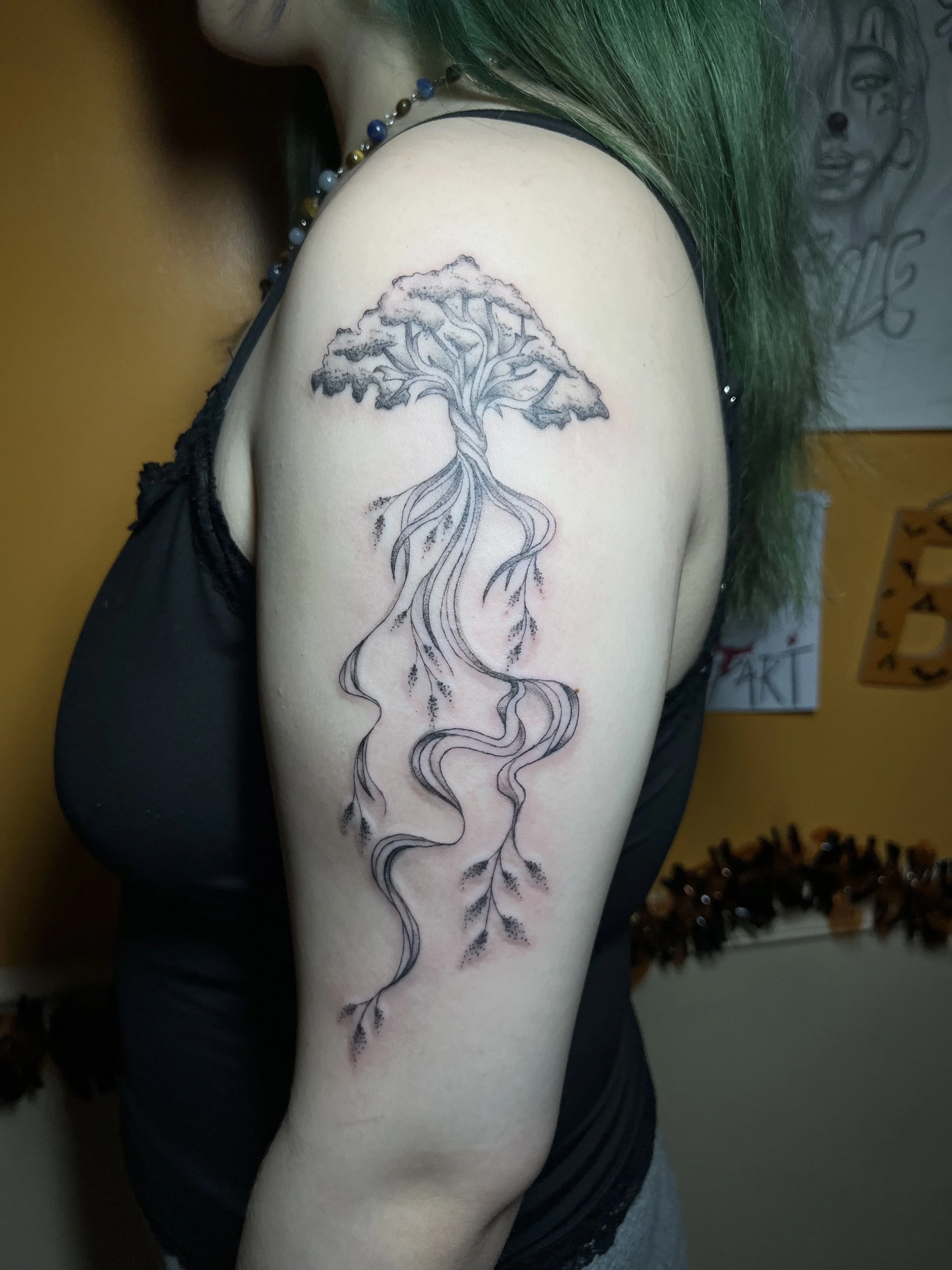Black and gray tattoo of a tree with roots and branches extending along the upper arm and shoulder, with detailed leaves, on a person with green hair wearing a black sleeveless top.