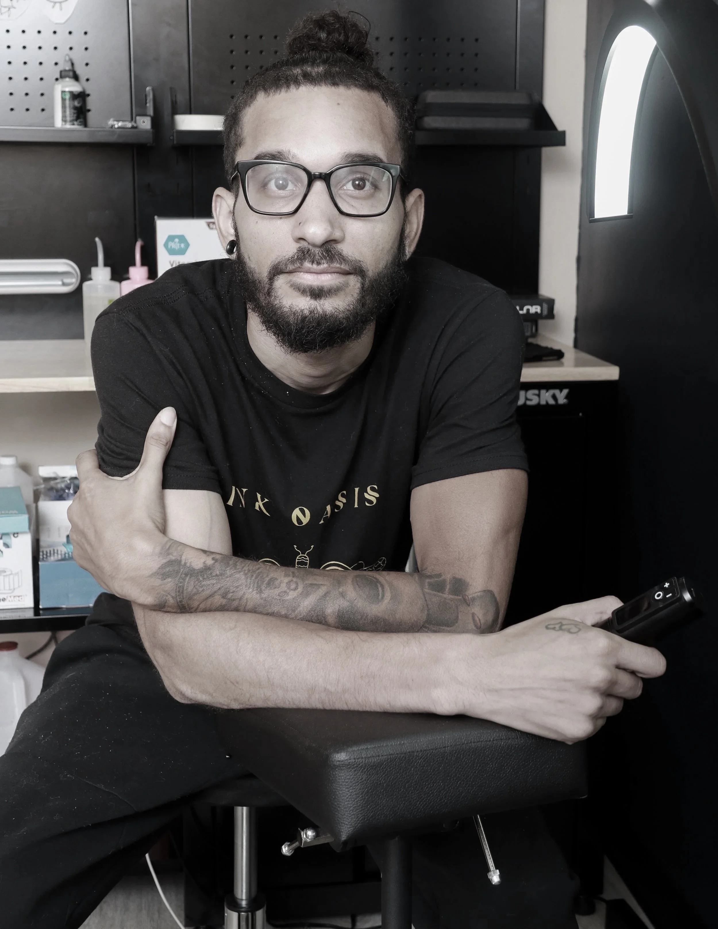 A man with glasses and a beard sitting on a tattoo chair, holding a tattoo machine, in a tattoo studio.