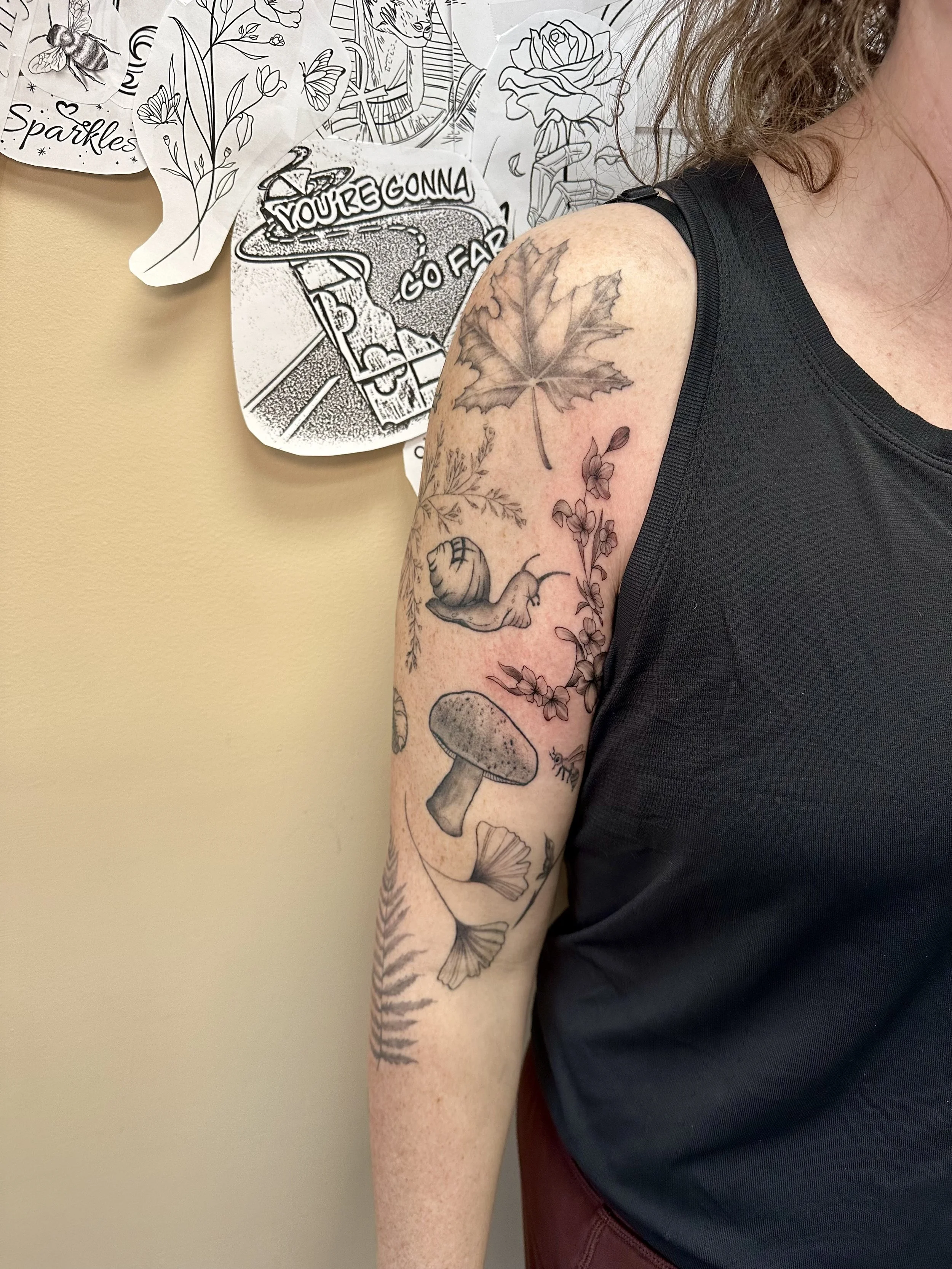 Tattoo of a large maple leaf, a small flower, a flying fish, a mushroom, on a person's upper arm.
