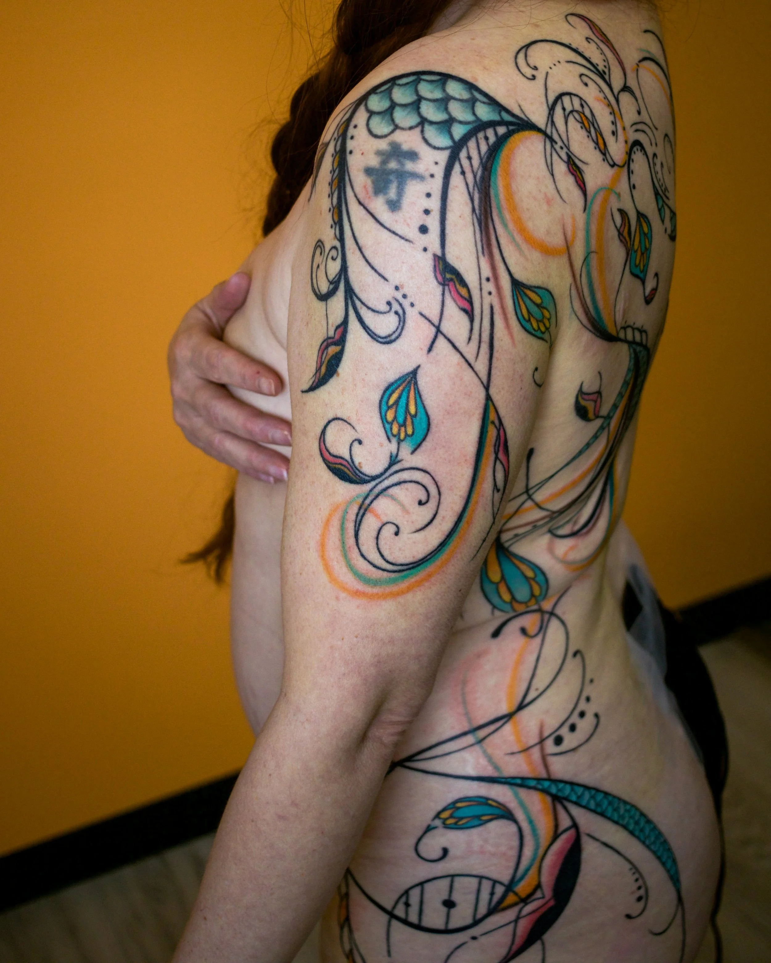 A person's arm and torso with colorful, intricate tattoos featuring floral and abstract designs, including a dragon, leaves, and swirling patterns on a yellow background.