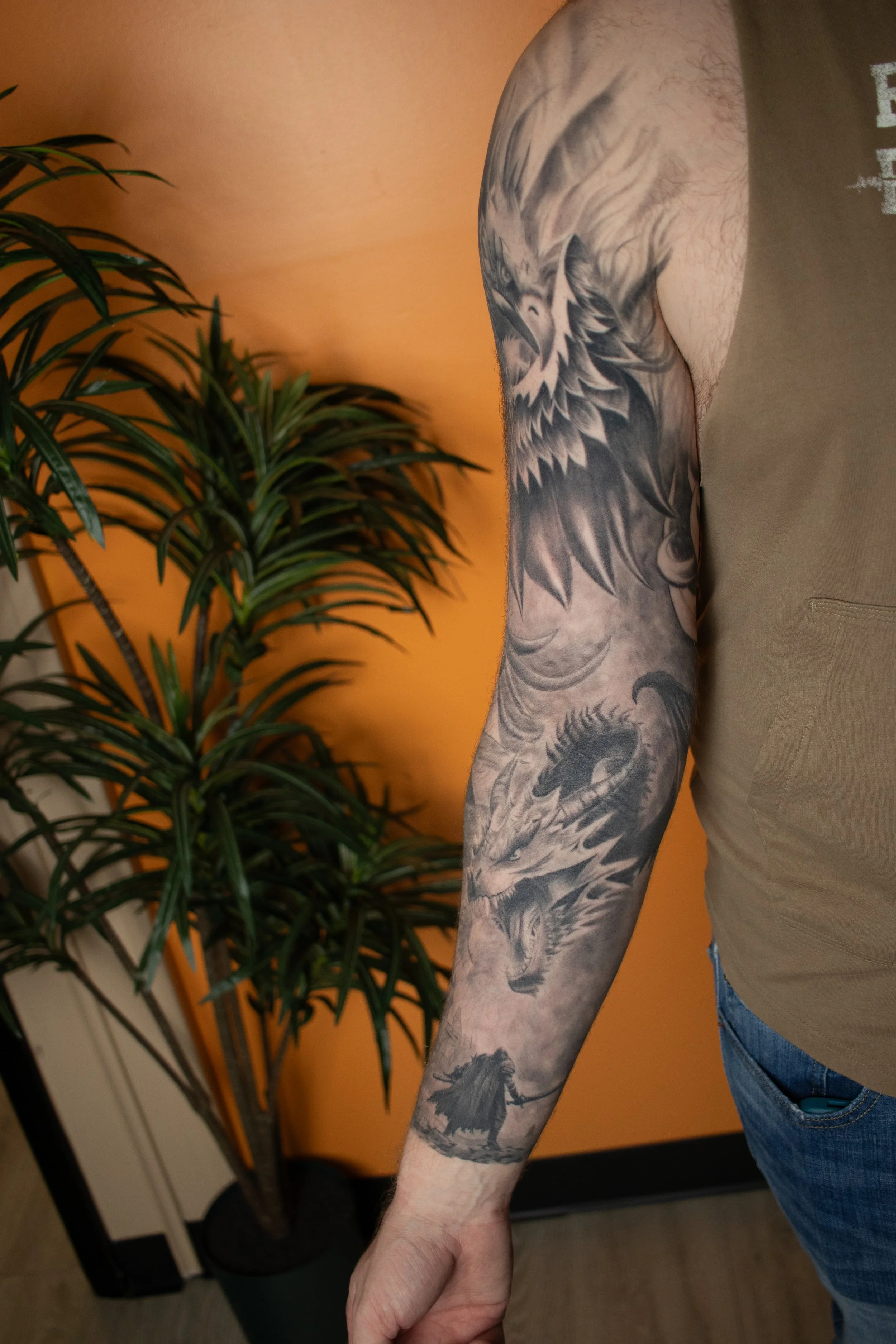 Close-up of a person's arm with a black and gray dragon tattoo, standing near a potted plant with long, green leaves, against an orange wall background.