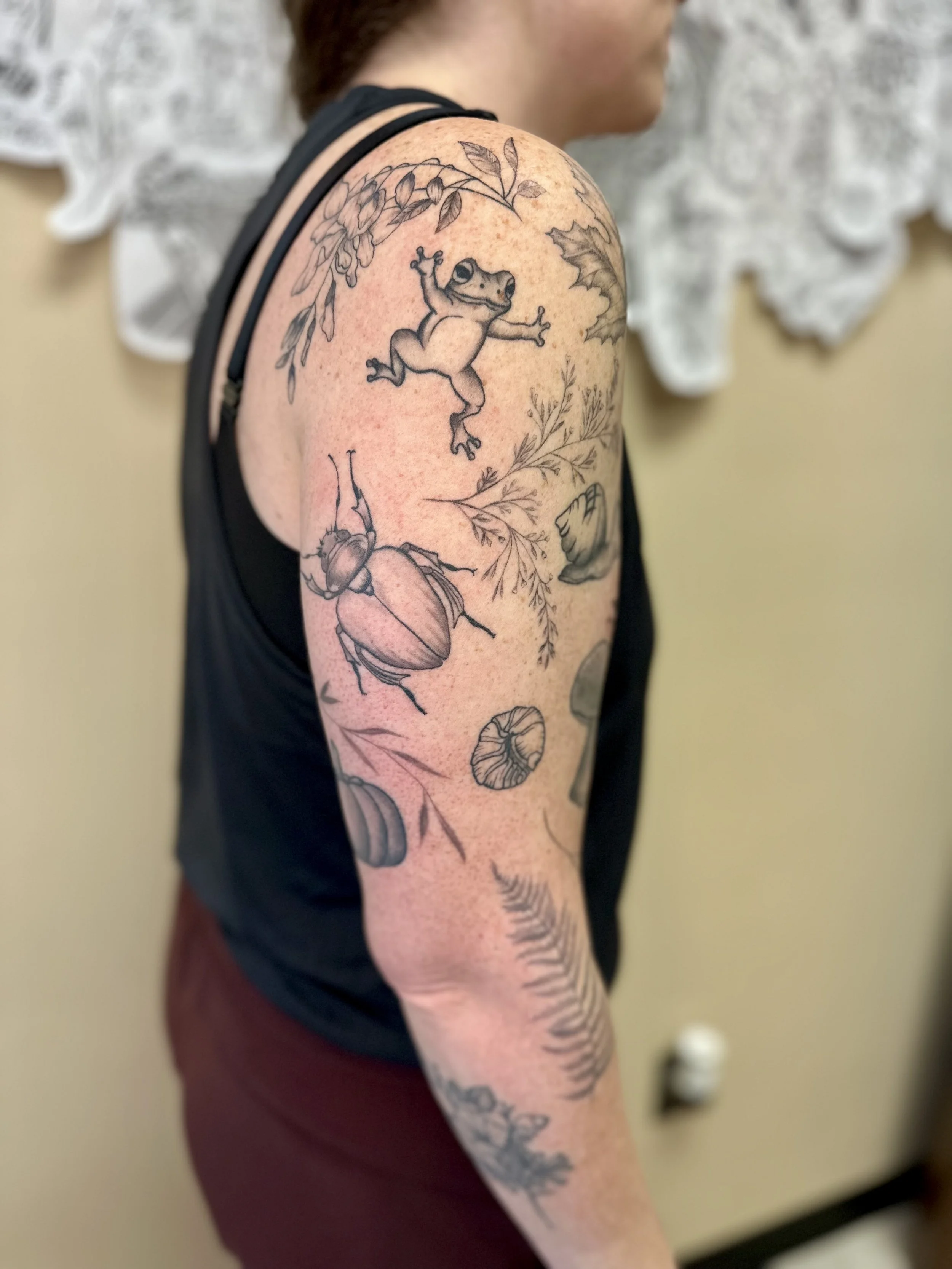 Tattoo sleeve on woman's upper arm featuring a frog, a beetle, leaves, mushrooms, and various plants in black ink.