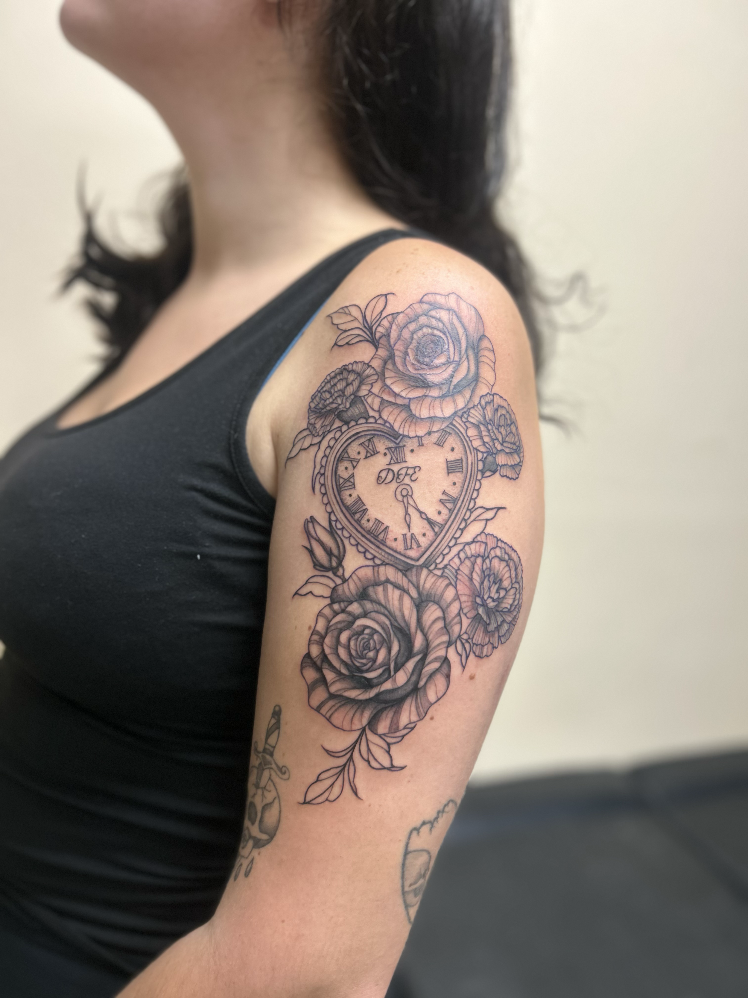 A woman's upper arm tattoo featuring a large, detailed rose, smaller flowers, and a clock with Roman numerals and the word 'DIE' across the center.