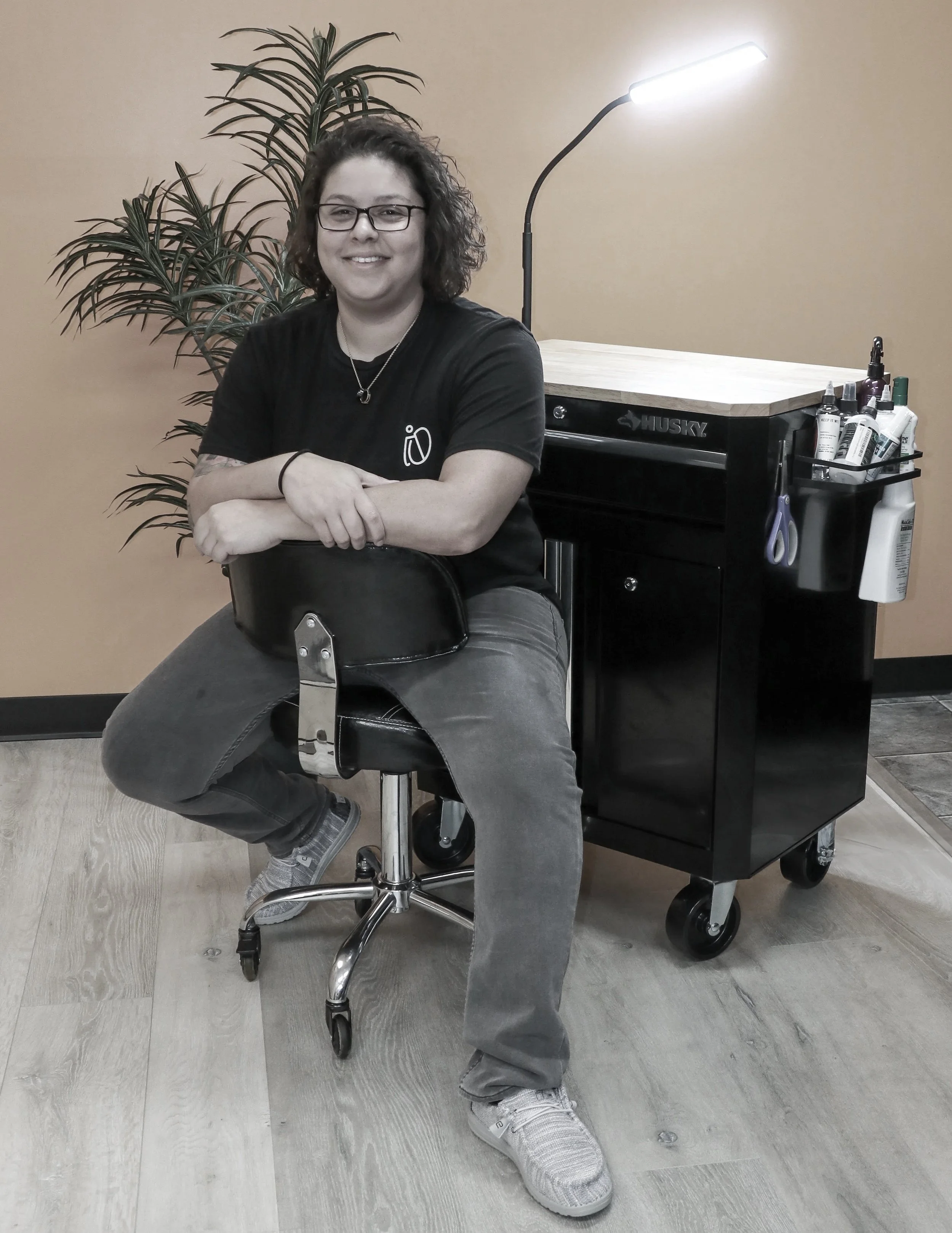 A smiling woman with curly hair, wearing glasses, a black t-shirt, and gray pants, sitting on a swivel stool in a salon or tattoo studio, with a rolling black cart holding various bottles and scissors, and a tall floor lamp, against a beige wall with a green potted plant in the background.