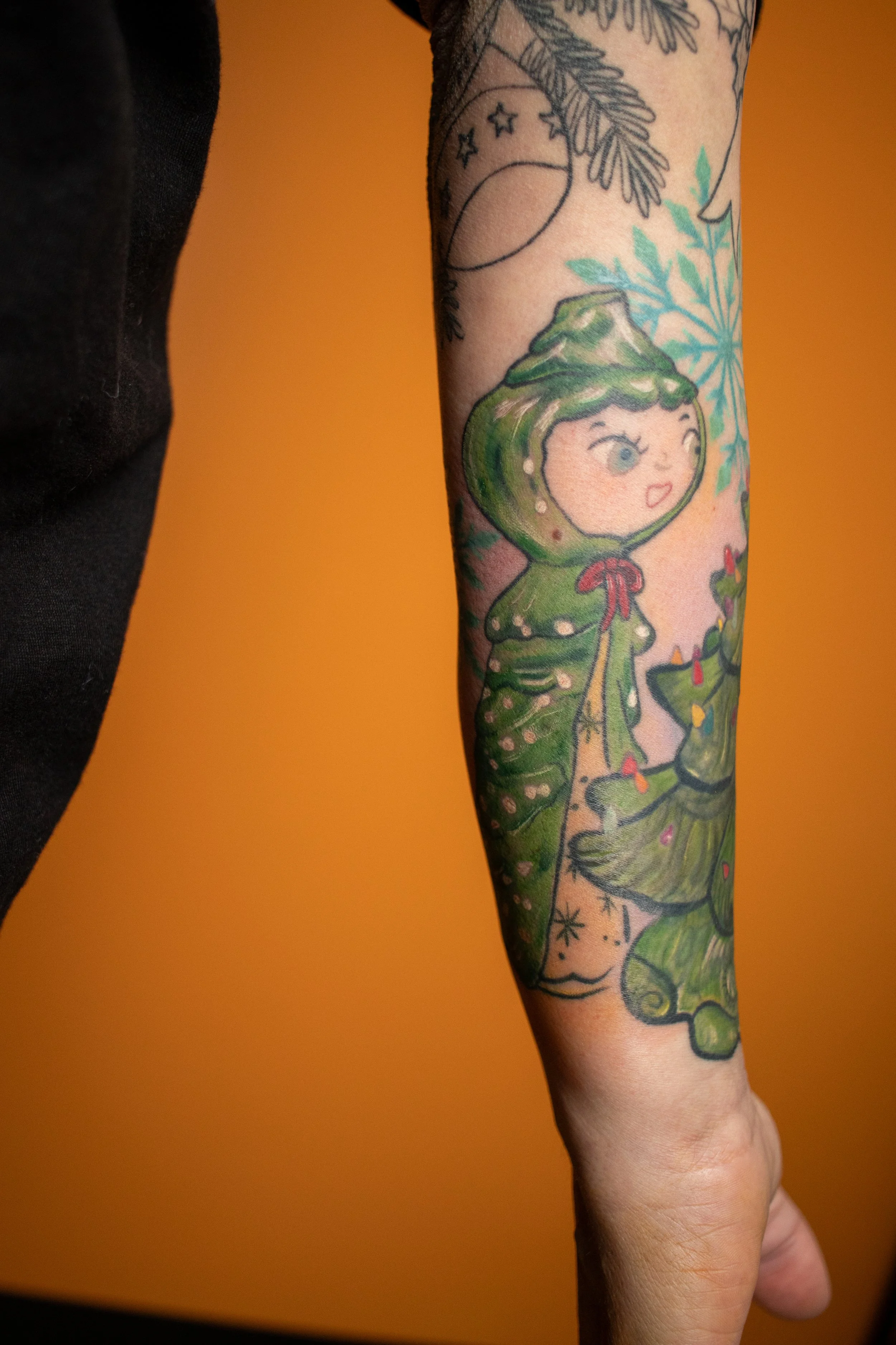 Colorful tattoo of a cartoon girl with green hair, wearing a hooded dress, on a person's arm with an orange background.