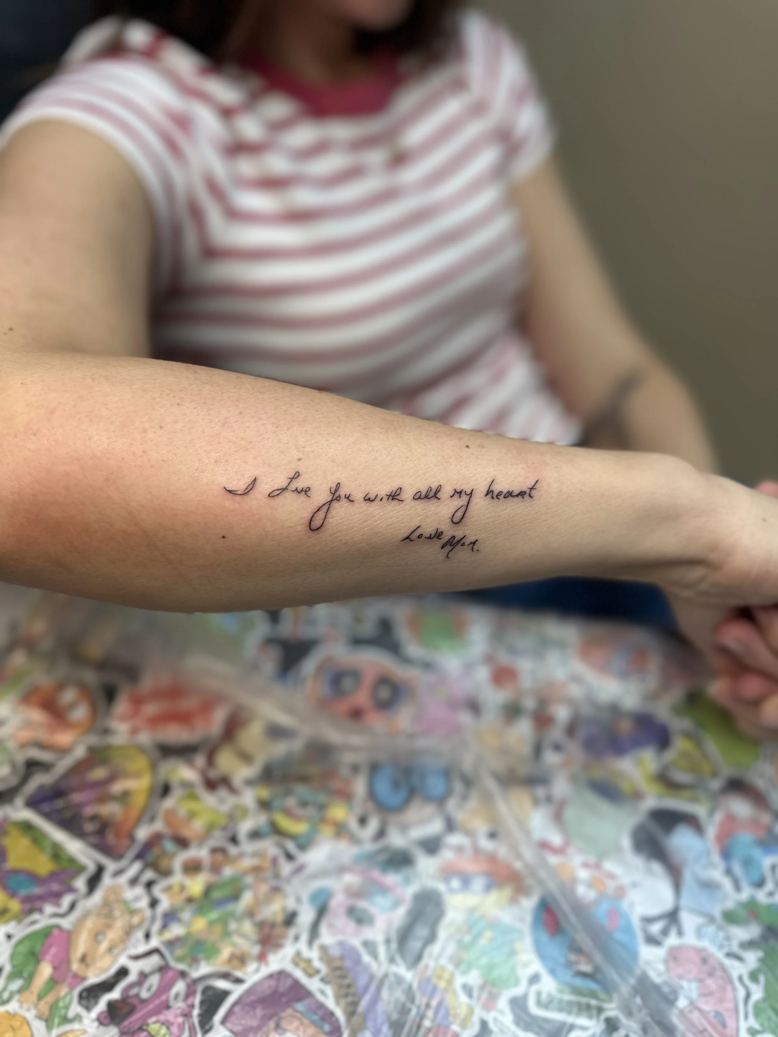 A person with short dark hair wearing a red and white striped shirt showing a tattoo on their forearm that reads "I Love You with all my heart. Love A.S." in cursive handwriting. The person is sitting at a table covered with colorful stickers.