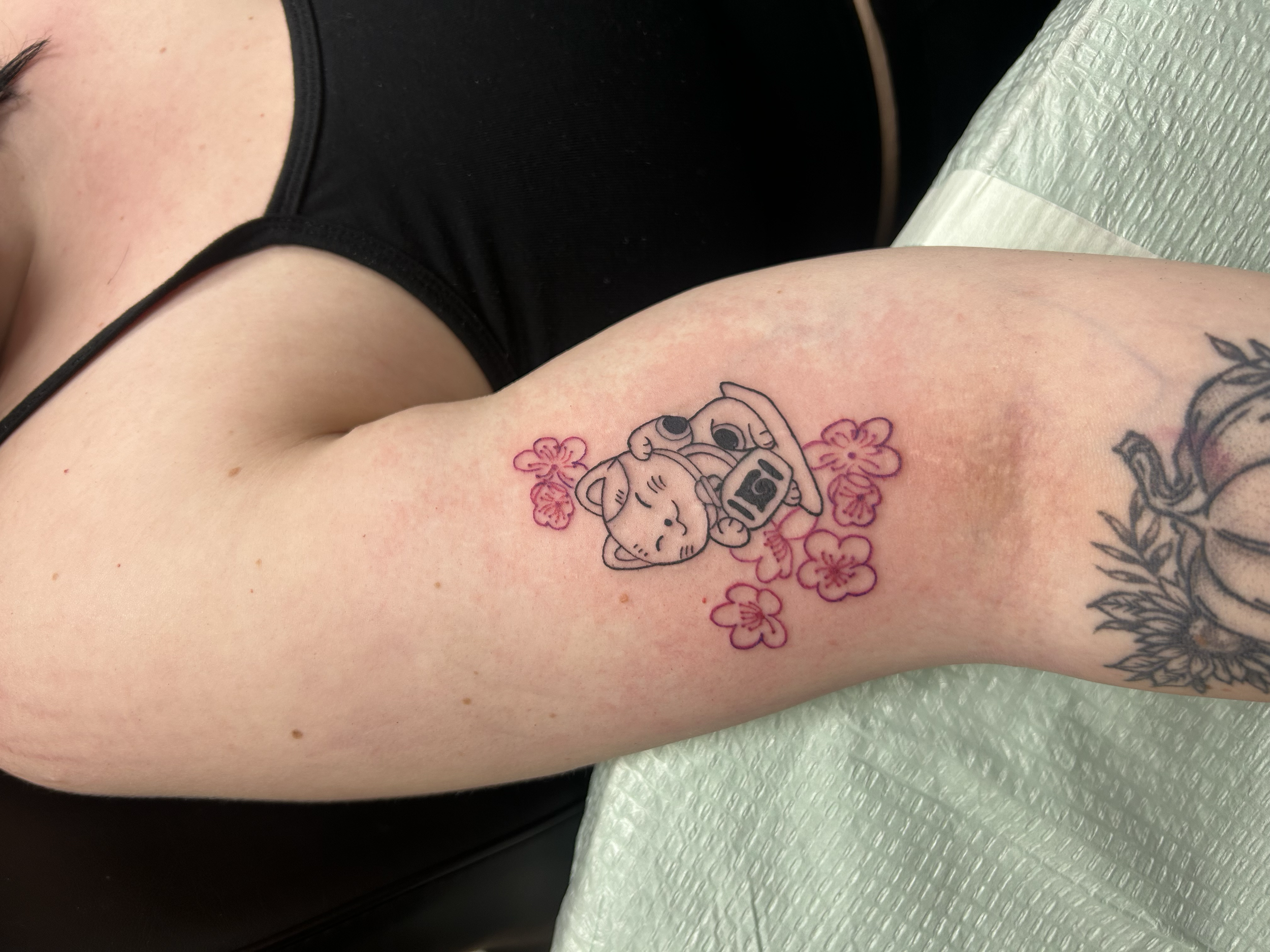 Tattoo of a cute cat holding a camera surrounded by pink flowers on person's upper arm.