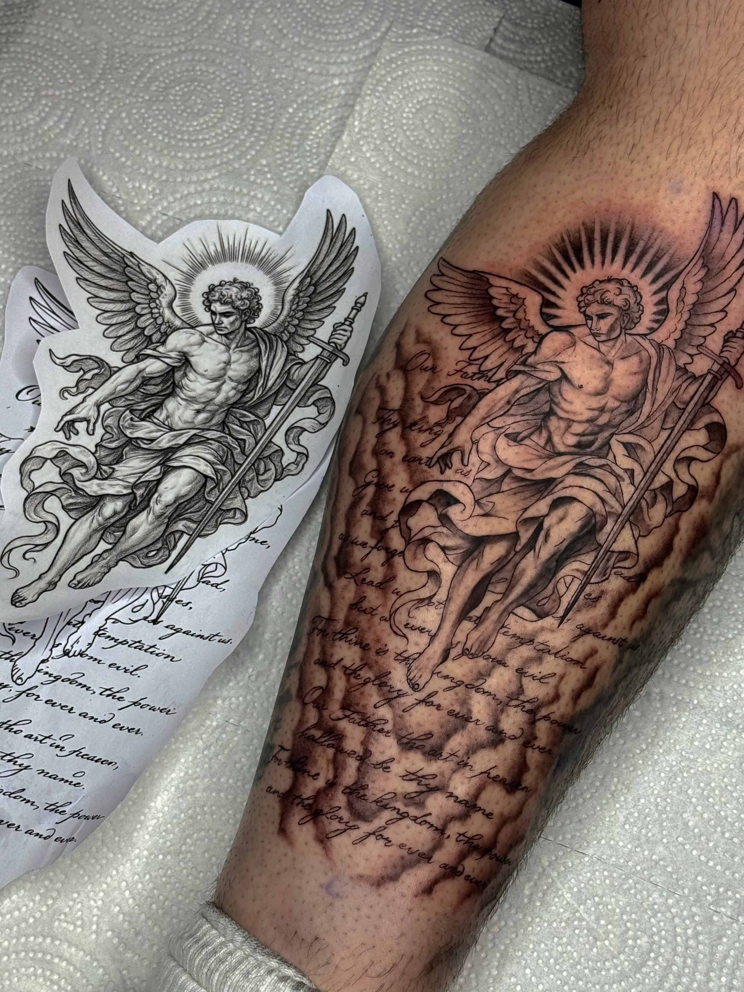 A tattoo of an angel with wings, holding a sword, with detailed shading and script writing, in progress on a person's arm.