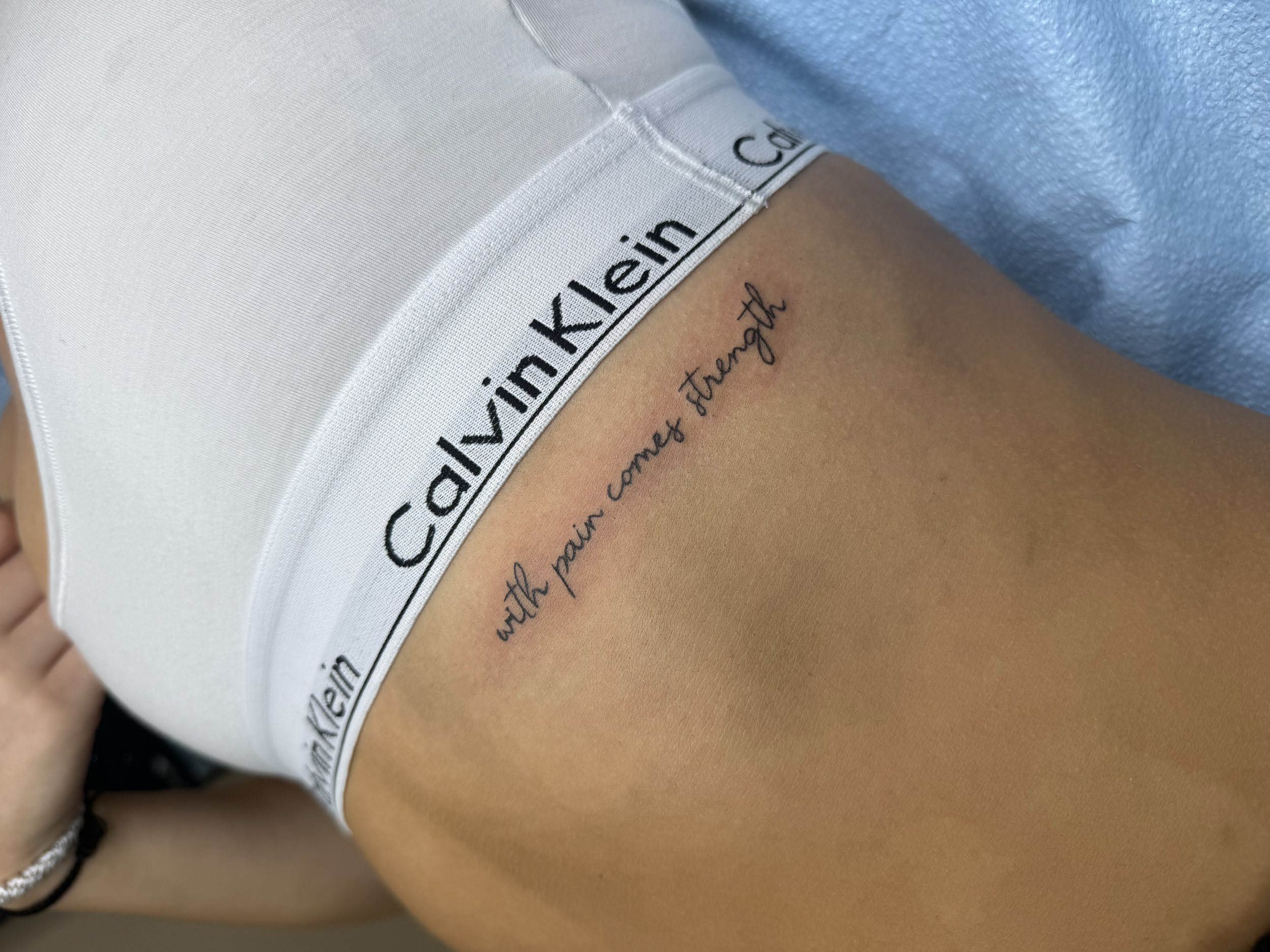 Close-up of a person's midsection wearing white Calvin Klein underwear with black text, revealing a tattoo that reads 'with pain comes strength' on the skin above the underwear