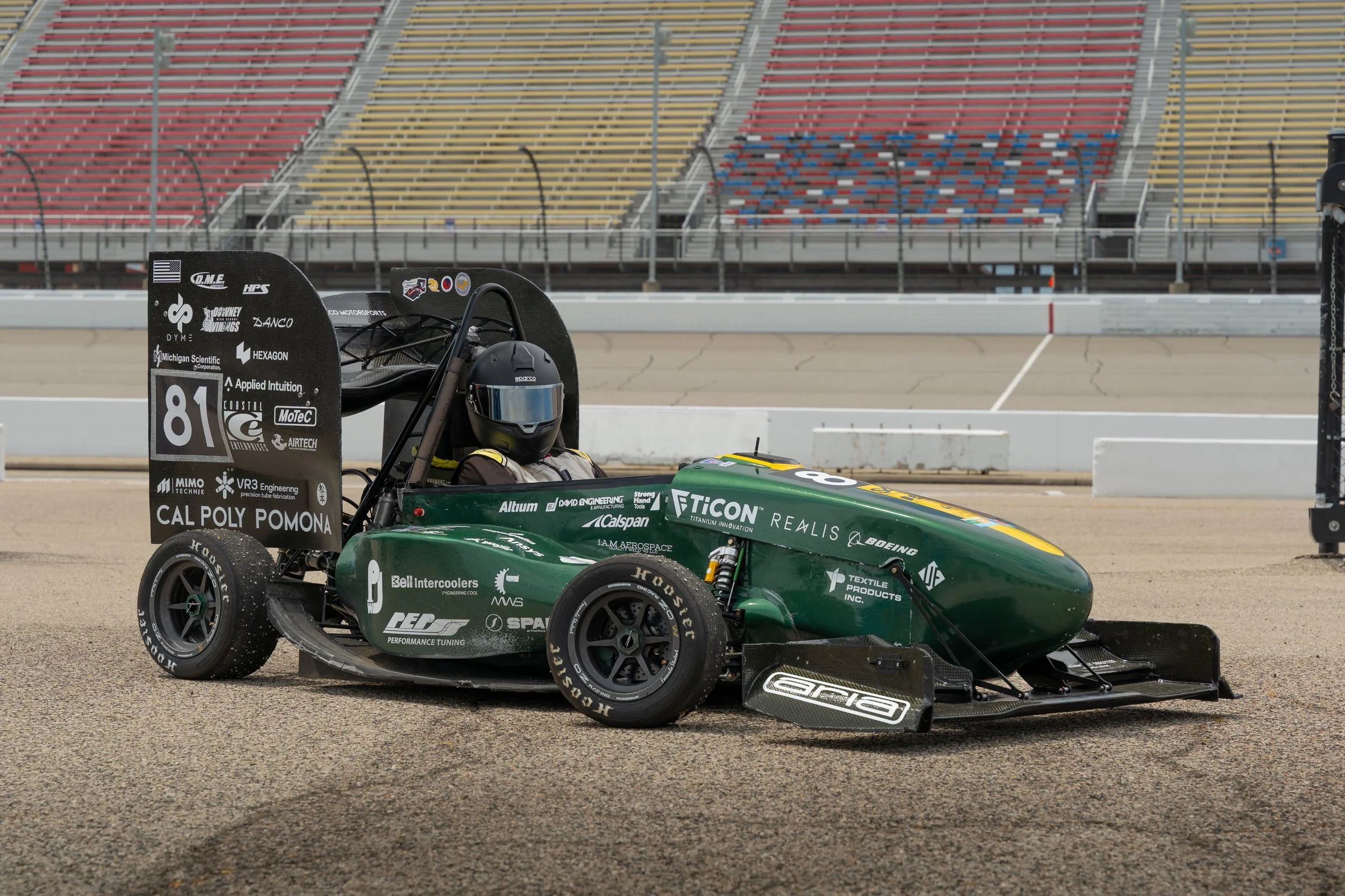 Green and black race car with the number 81 on the rear wing, parked on a racetrack with empty grandstands in the background.
