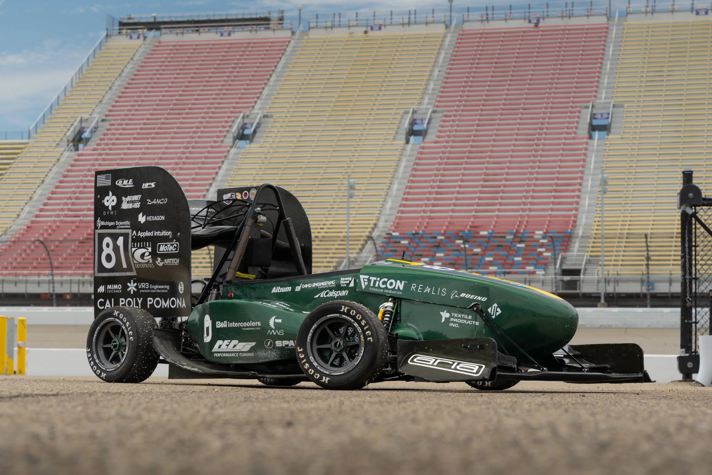 Green and black race car with the number 89 on the rear wing, parked on a racetrack with empty grandstands in the background.