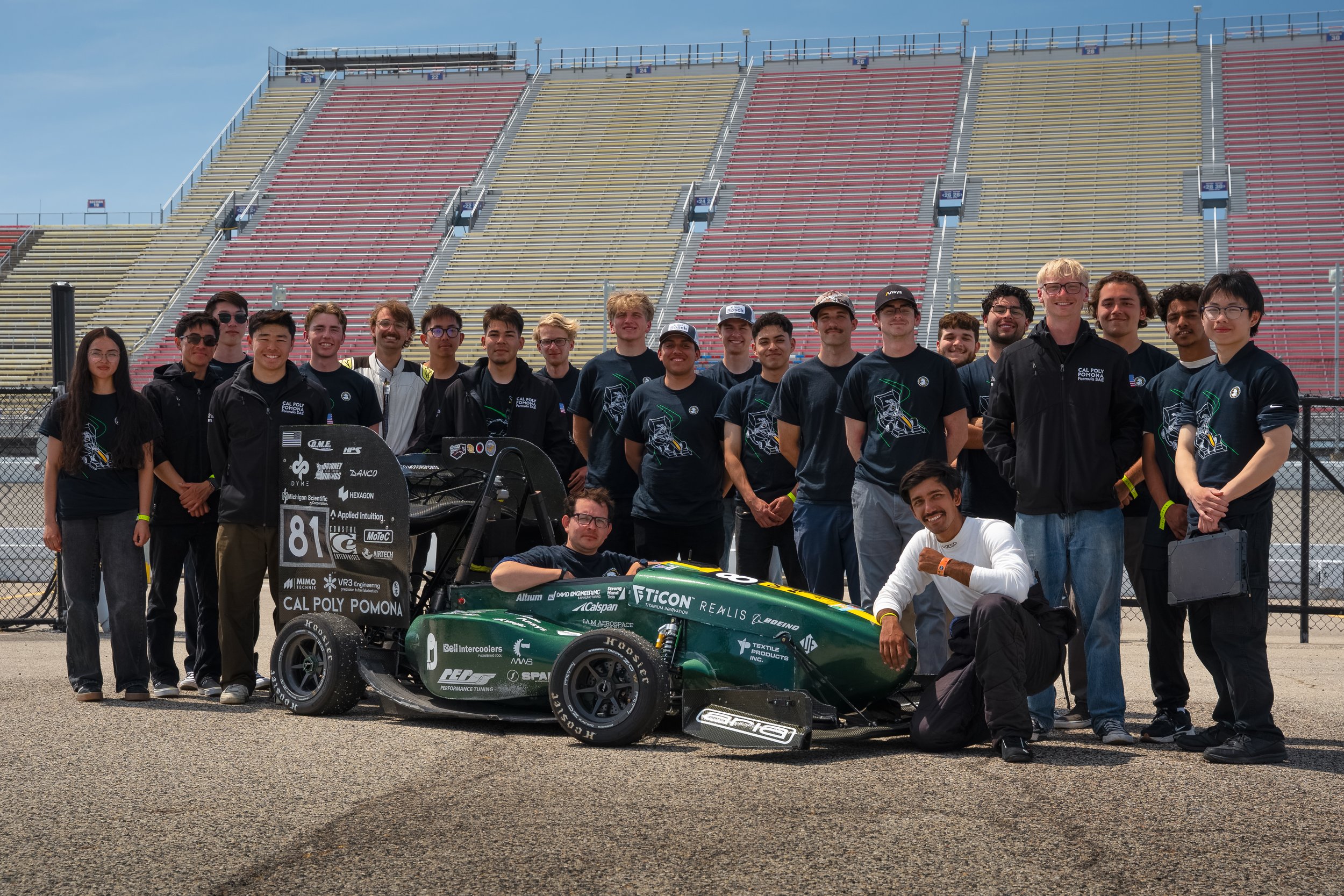 A group of young people, including racing drivers, standing on a racetrack behind a green race car. The group includes individuals in racing suits and casual clothing, some wearing helmets and hats. The background features empty bleachers and a chain-link fence.