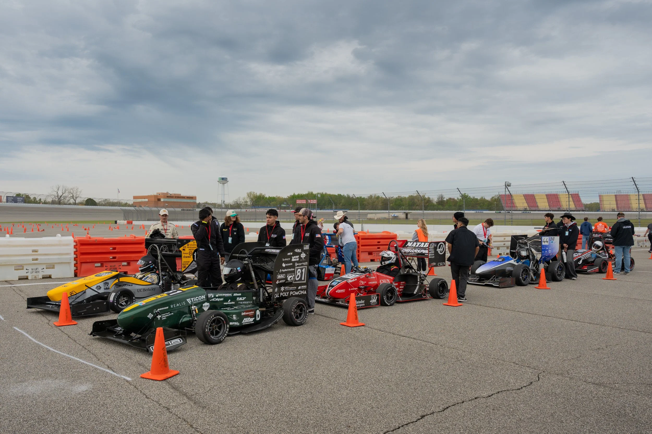Race cars and teams preparing at a race trackpit area under cloudy skies, with people working on vehicles and socializing.