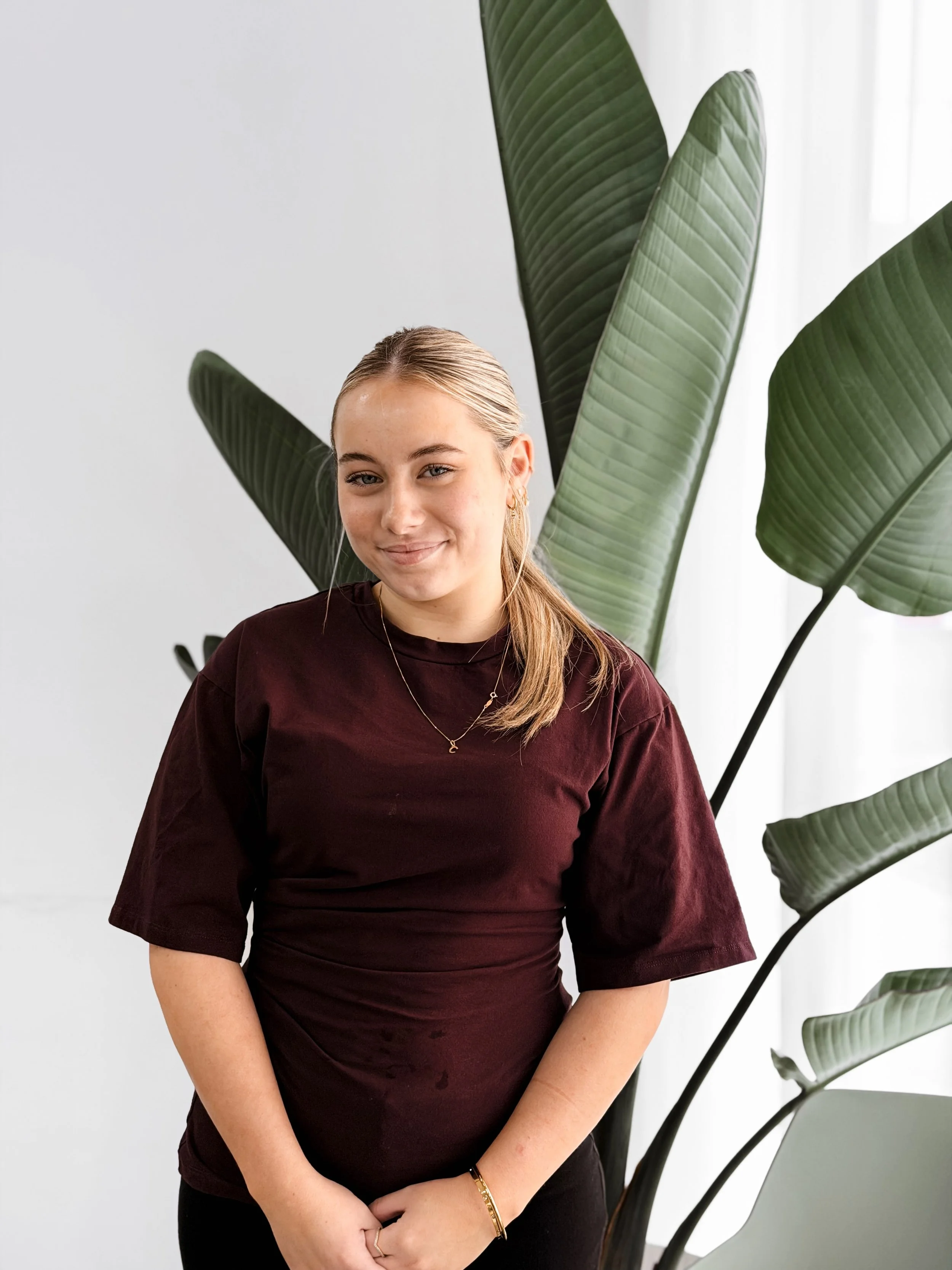 A woman with blonde hair tied back, wearing a maroon t-shirt, gold jewelry, and a subtle smile, standing in front of large green plant leaves and a white wall.