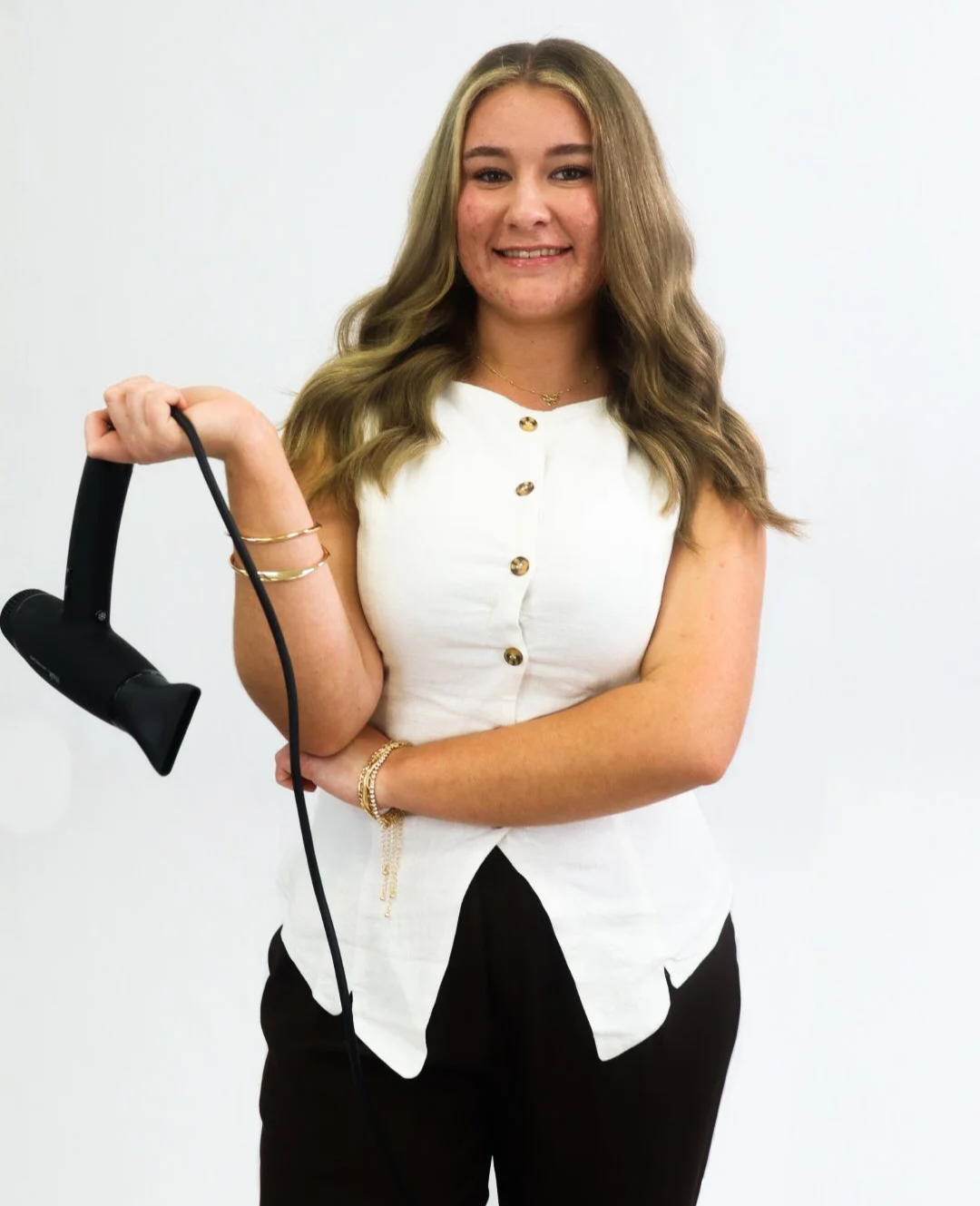 A woman with long blonde hair smiling, wearing a sleeveless white button-up top, holding a black hair steamer, standing against a white background.
