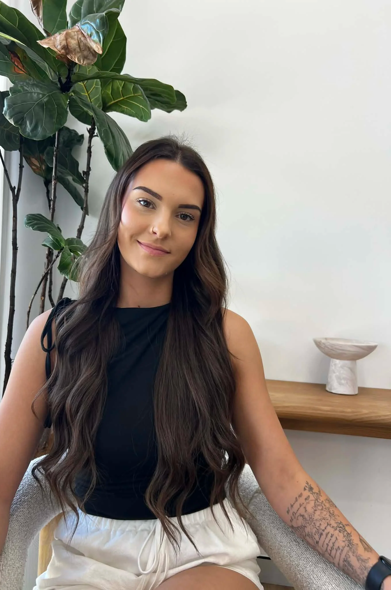 A young woman with long, wavy brown hair and tattoos on her arm, smiling and sitting in a cozy, modern space with a plant and decorative bowl in the background.