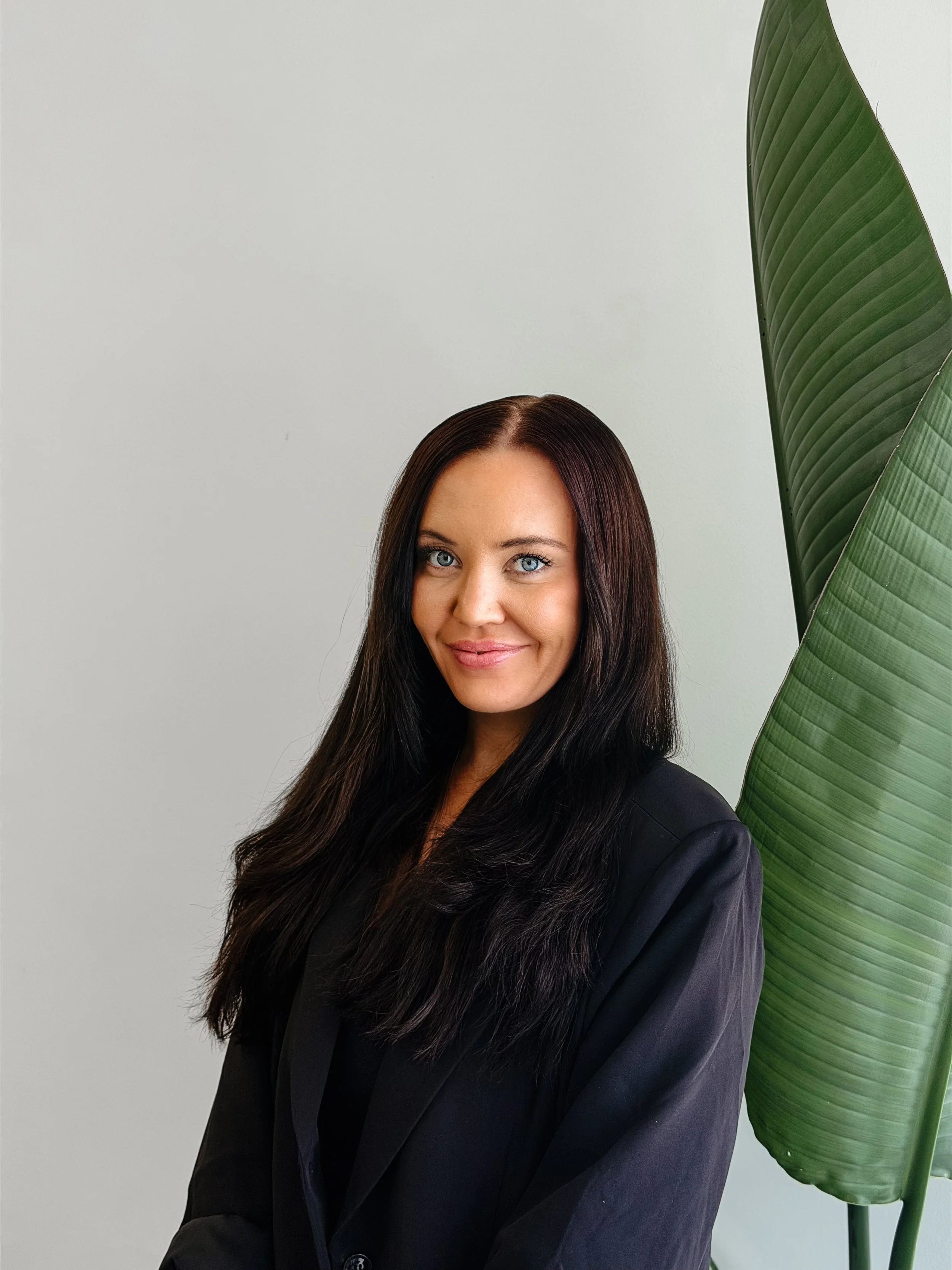 A woman with long dark hair and blue eyes smiling, wearing a black blazer, standing in front of a plain light-colored wall with a large green tropical leaf on her right side.