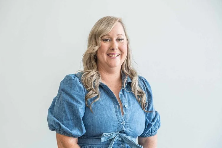 A woman with long, wavy blonde hair wearing a blue denim blouse with puffed sleeves, smiling and standing against a plain white background.