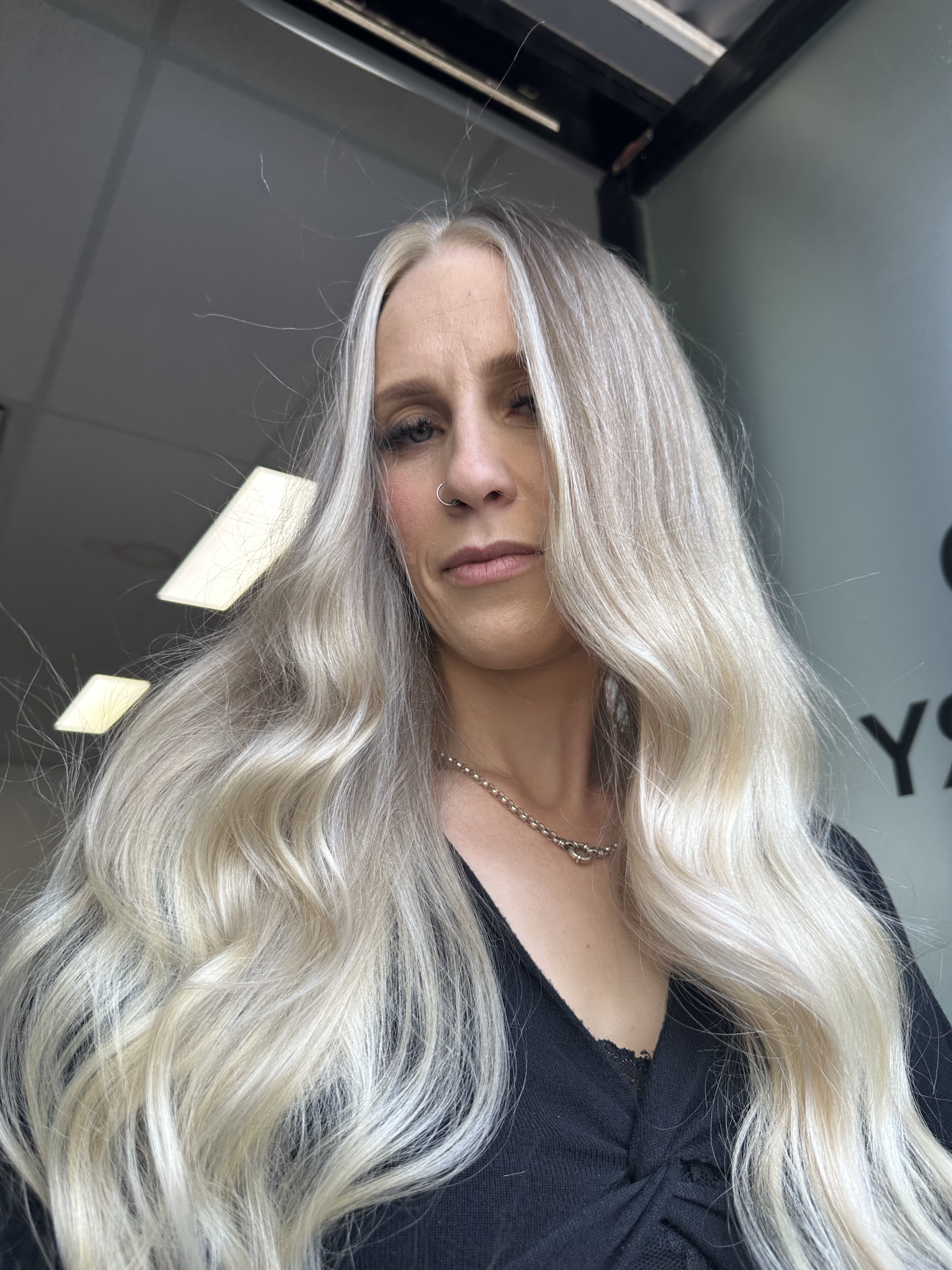 A woman with long, wavy platinum blonde hair and light skin takes a selfie in a room with gray walls and a black ceiling. She has a small nose ring, wears a silver chain necklace, and has a slight smile with one eye slightly closed.