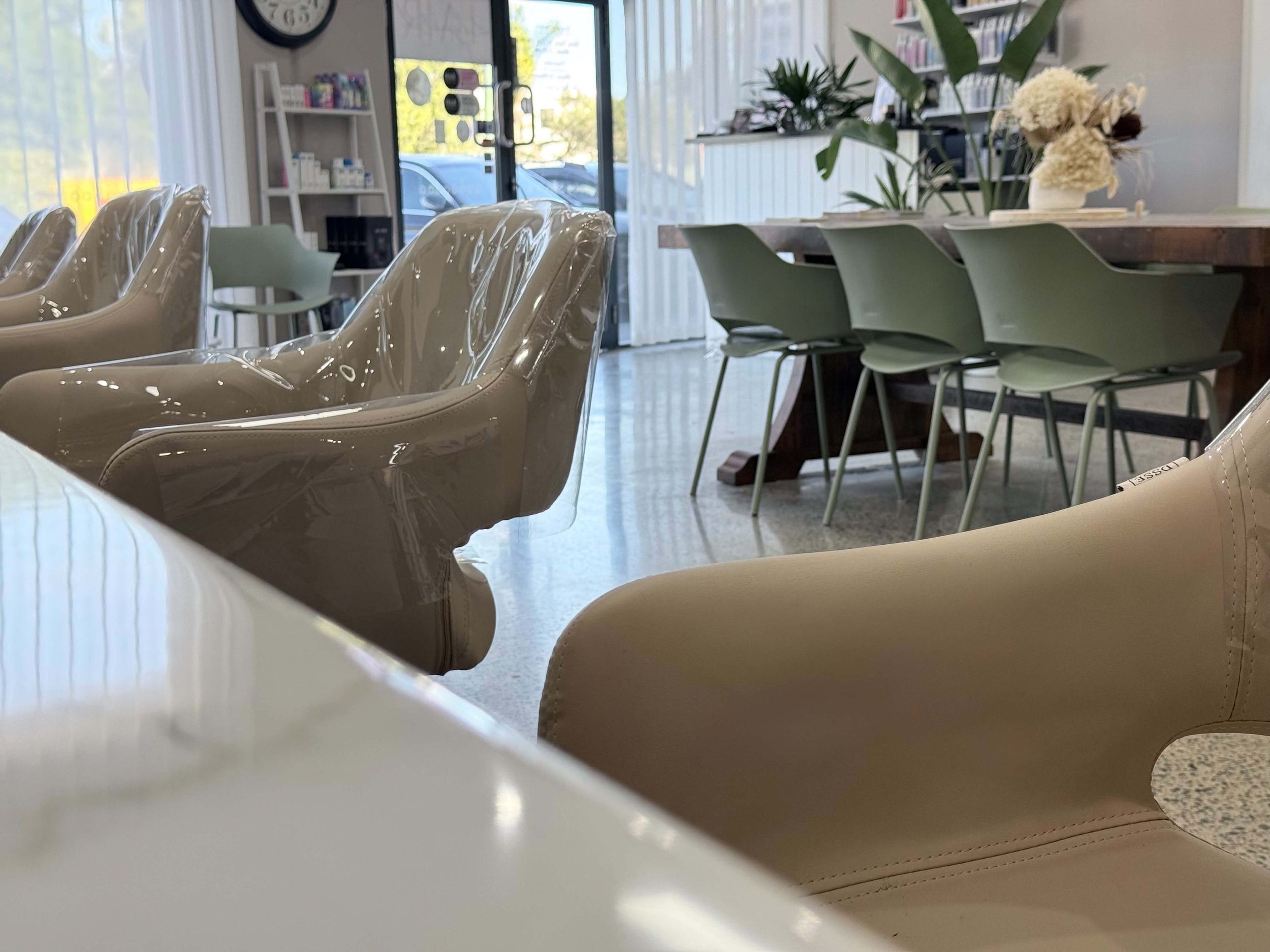 Interior of a waiting area or lobby with chairs covered in plastic, a long wooden table with green chairs, and plants on the table and in the background, near the window.