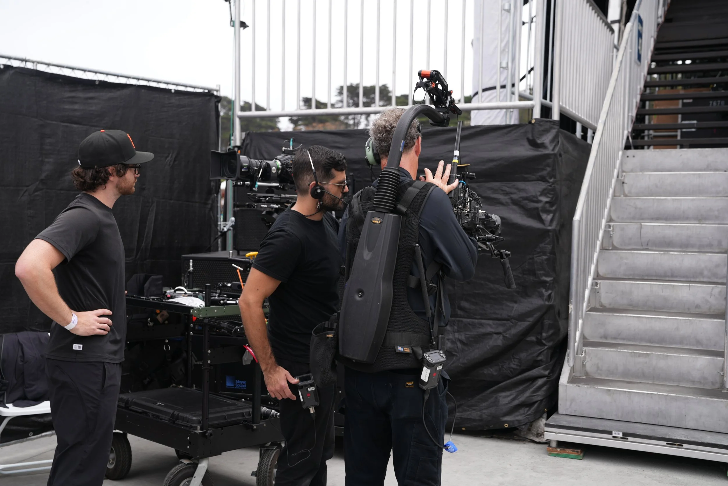 Three crew members operating professional video camera equipment at an outdoor event.