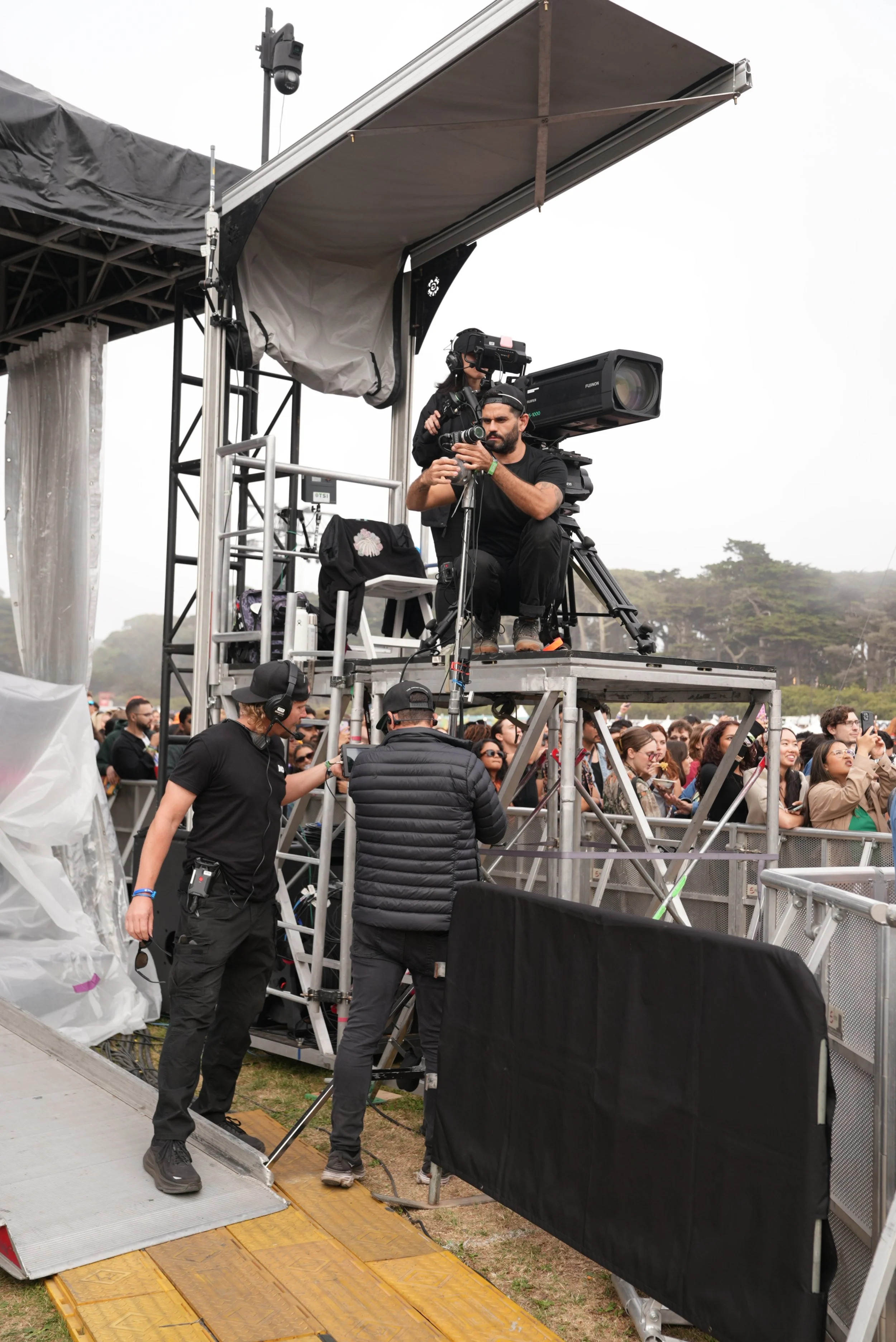Photographers and camera operators filming at an outdoor event with a crowd of spectators.
